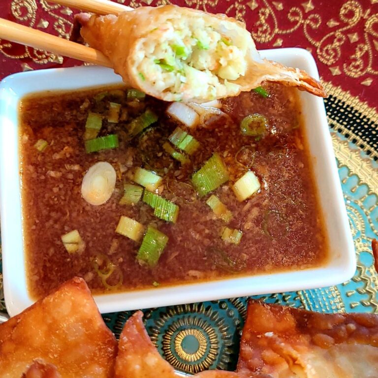 A fried shrimp wonton that is cut in half to show the shrimp filling held by chop sticks over a soy-based dipping sauce.
