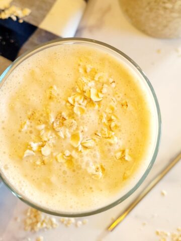 Smoothie in a glass on a counter top with oats on top, a straw, and a kitchen towel.