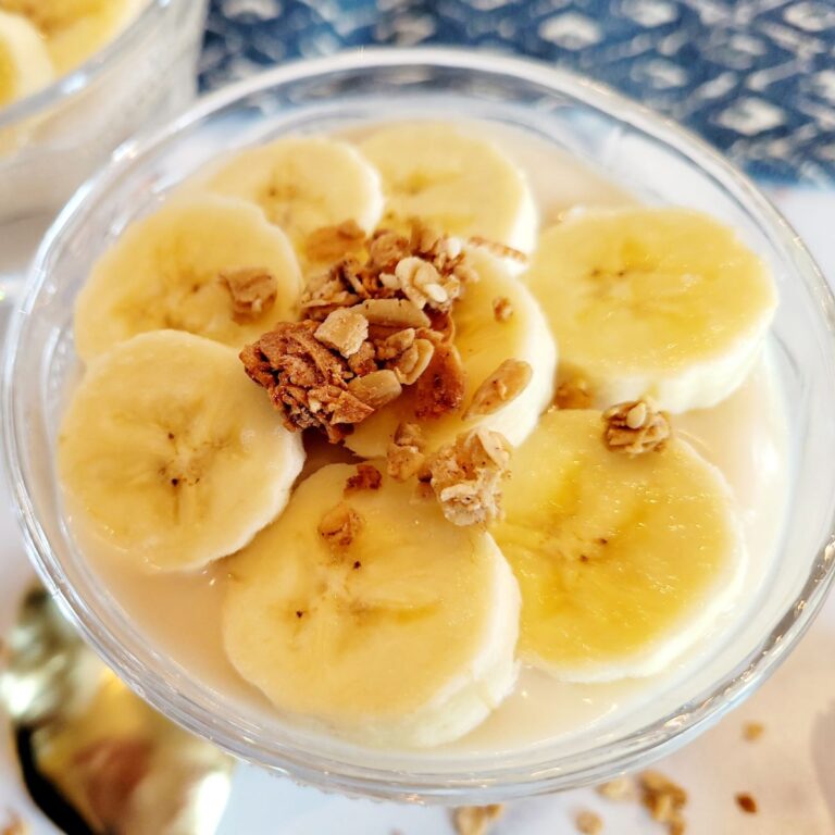 Pudding in ramekin with sliced bananas and granola on top surrounded by a spoon and kitchen towel.
