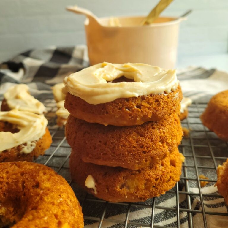 Stack of banana donuts with frosting on a counter top surrounded by more donuts and a mixing bowl.
