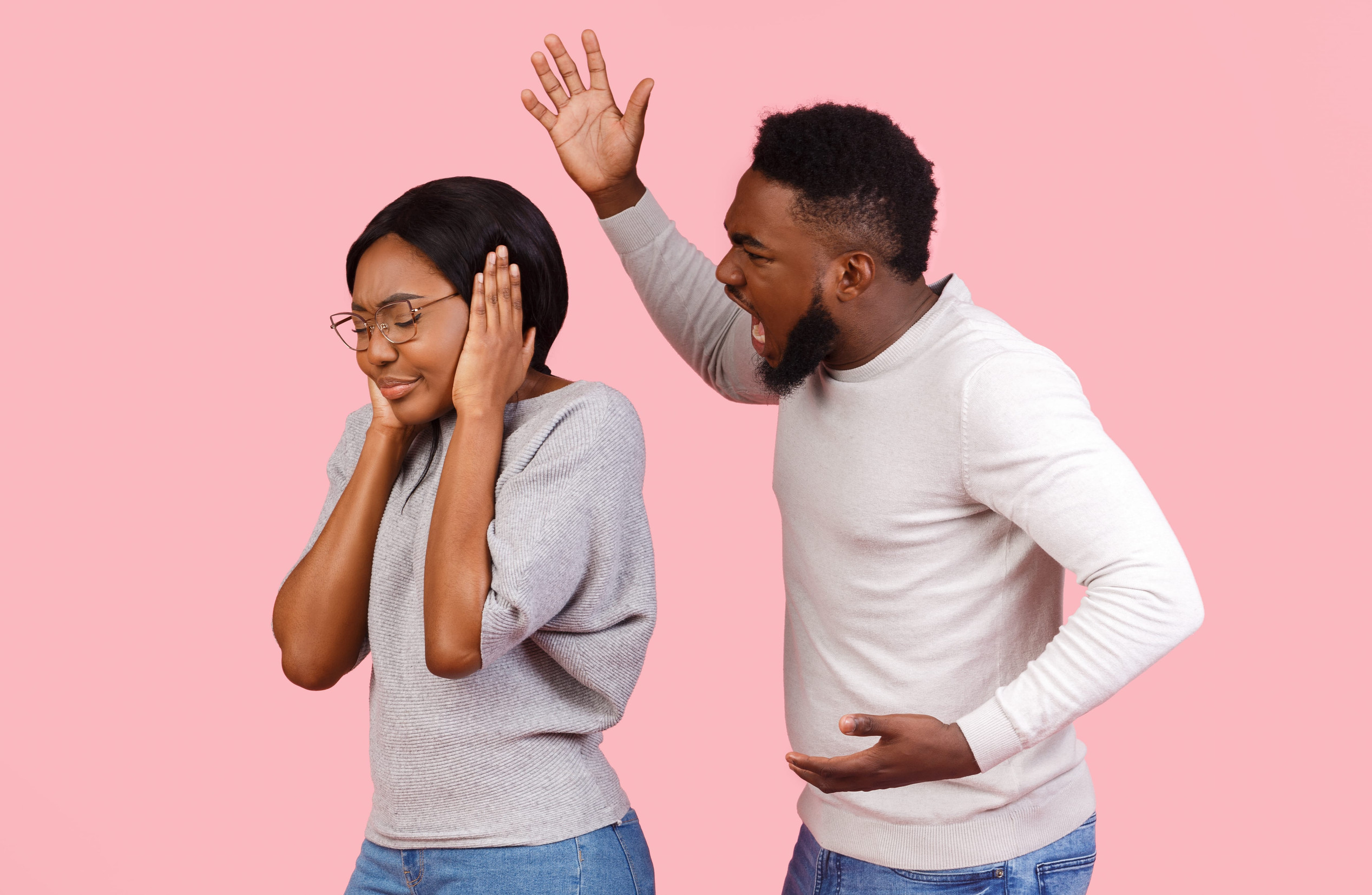 Domestic violence. Young african american man shouting at his girlfriend, woman covering ears with hands, pink studio background