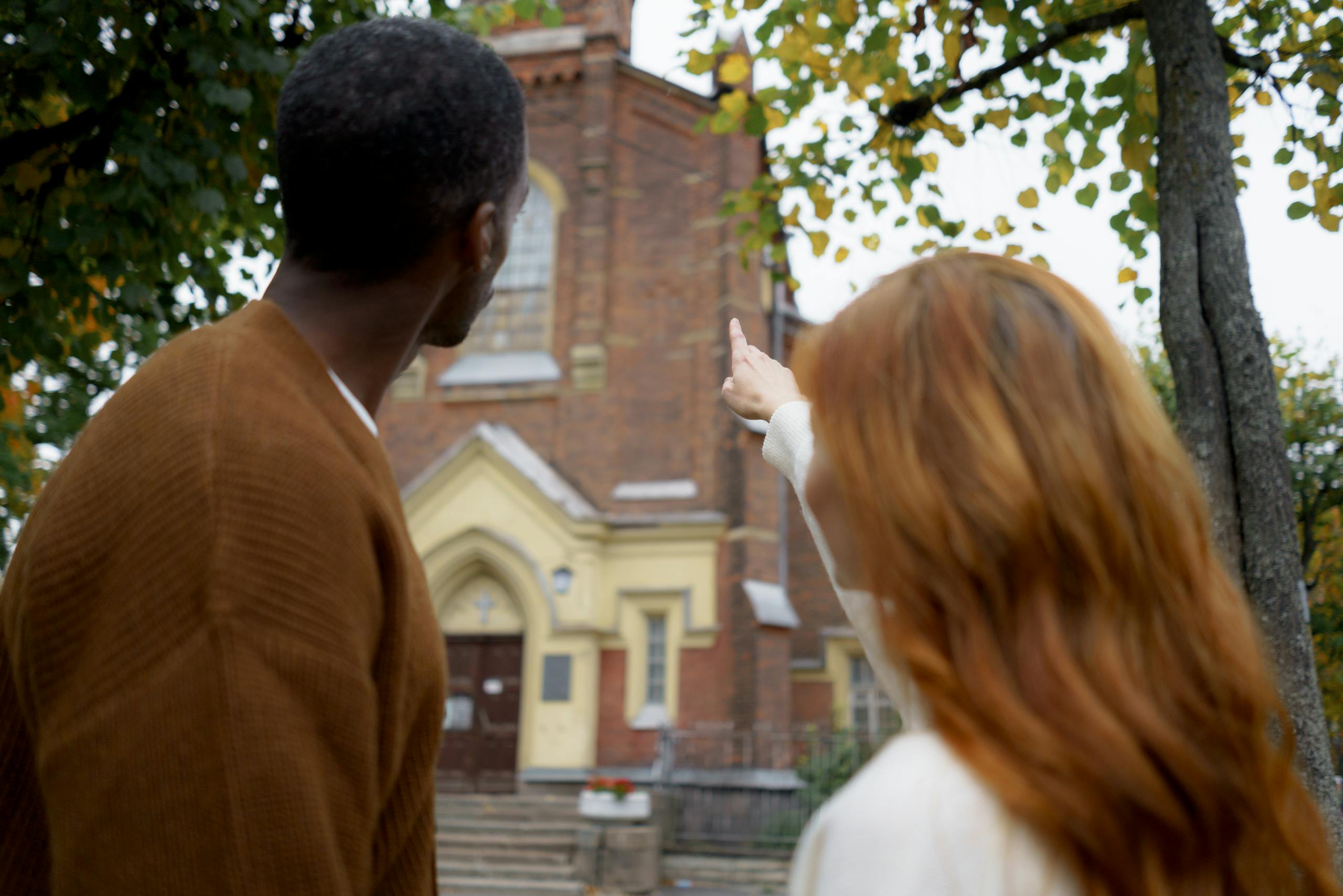 A black man and a caucasian woman, seen from behind, pointing at a historic church facade outdoors.