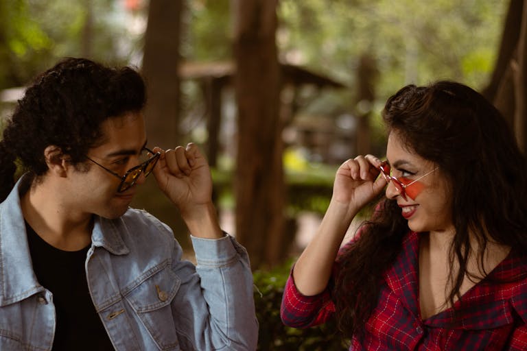 A cheerful couple enjoys a playful moment outdoors adjusting their sunglasses.