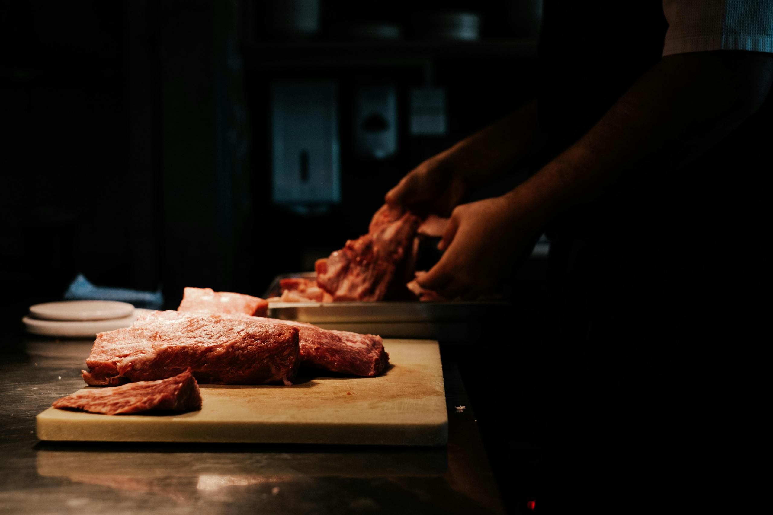 A chef skillfully cuts raw pork on a wooden board in a dimly lit kitchen, emphasizing culinary artistry.