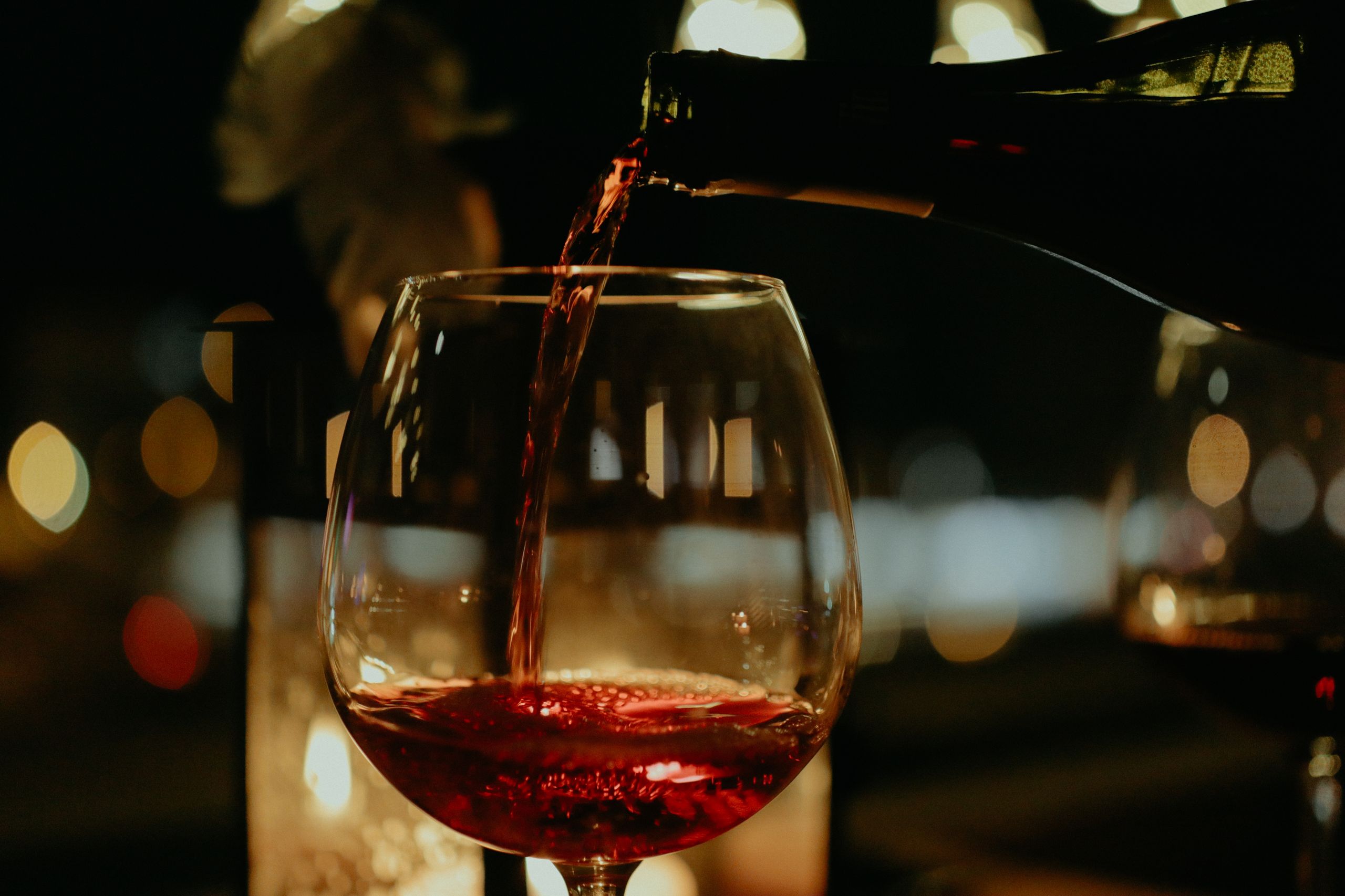 A close-up view of red wine being poured into a glass with blurred lights in the background.
