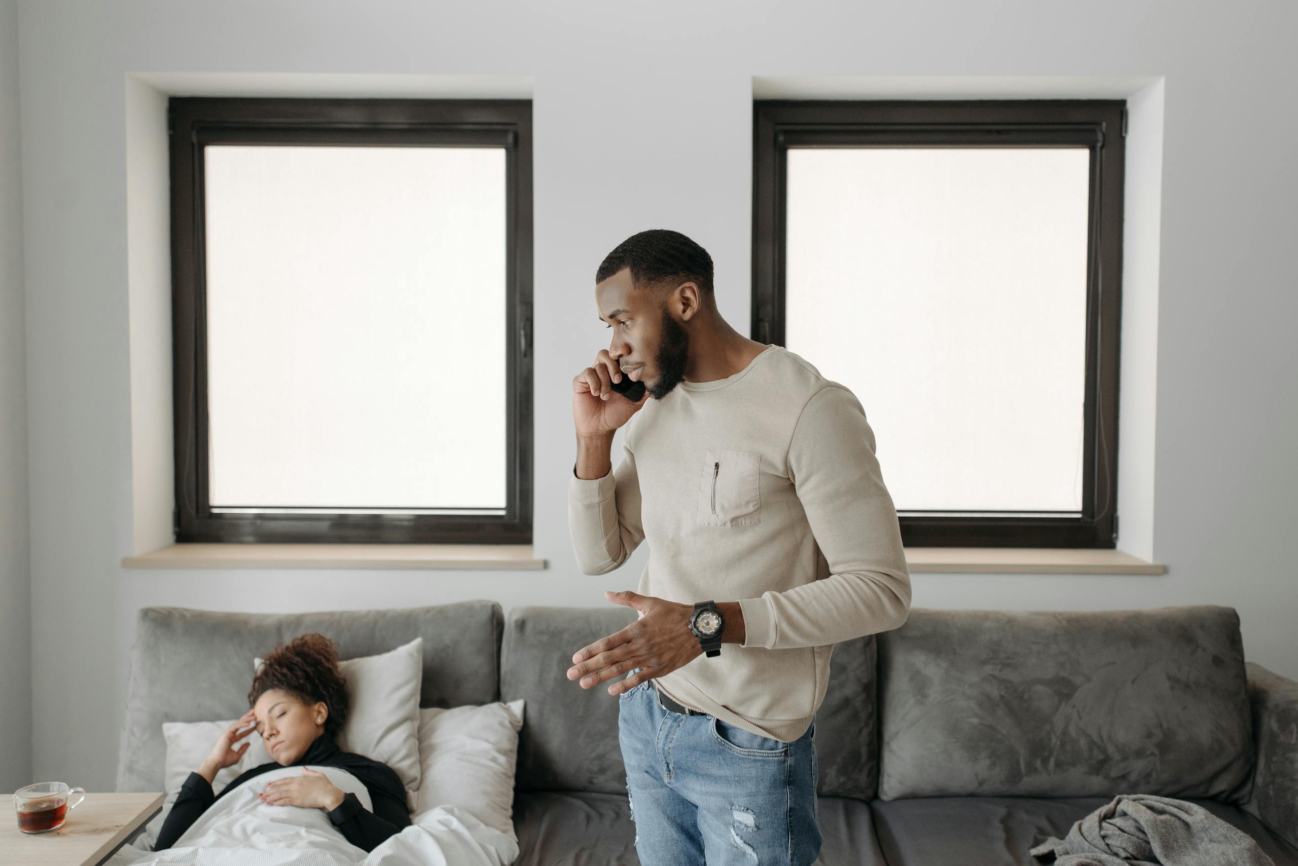 A concerned man makes a phone call while a woman sleeps unwell in bed, indicating urgency and care.