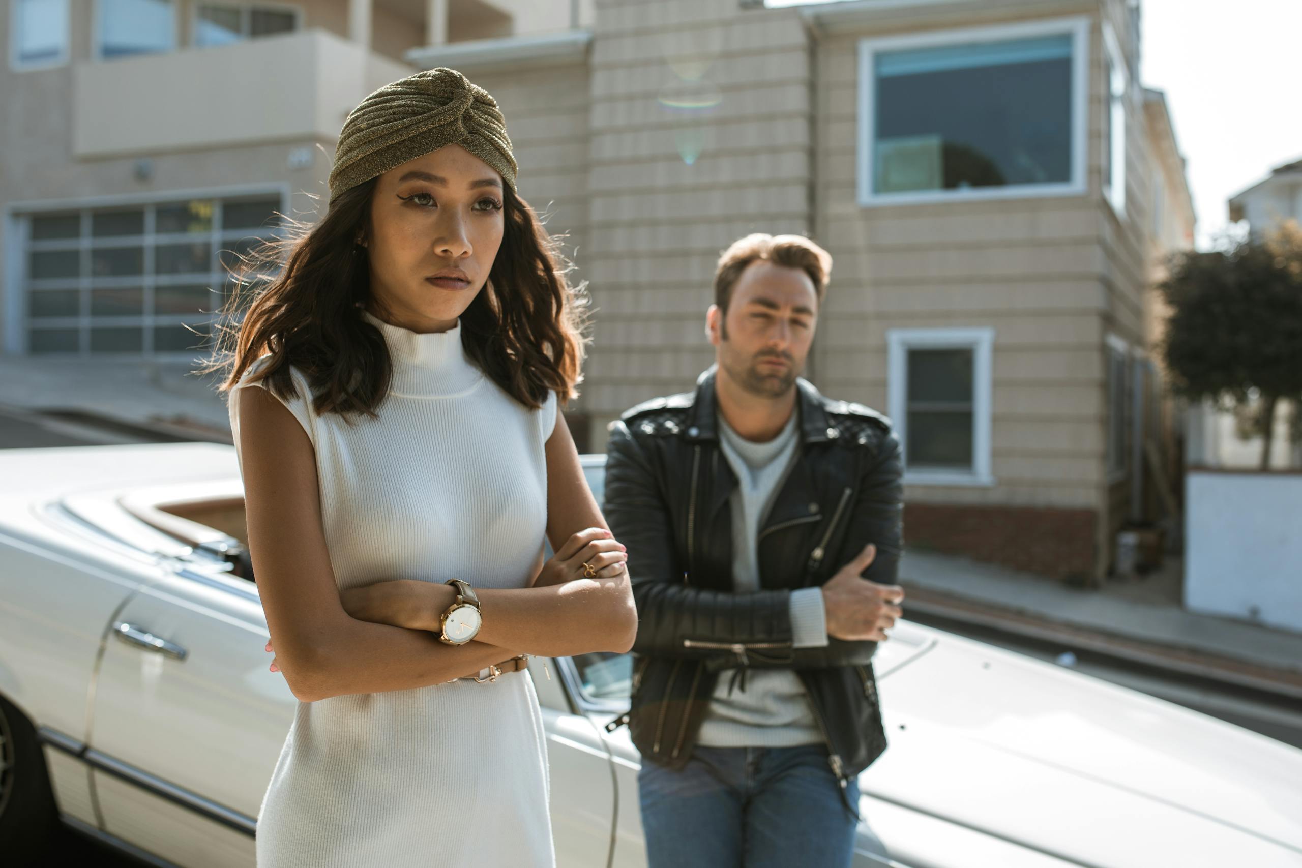 A couple appears upset, standing apart on a sunny urban street near a vintage car.