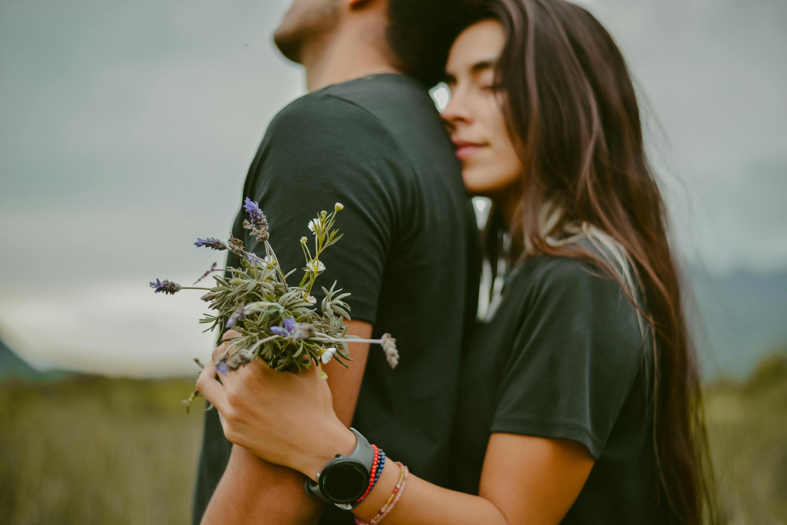 A couple embraces with love in a serene outdoor setting, holding flowers.