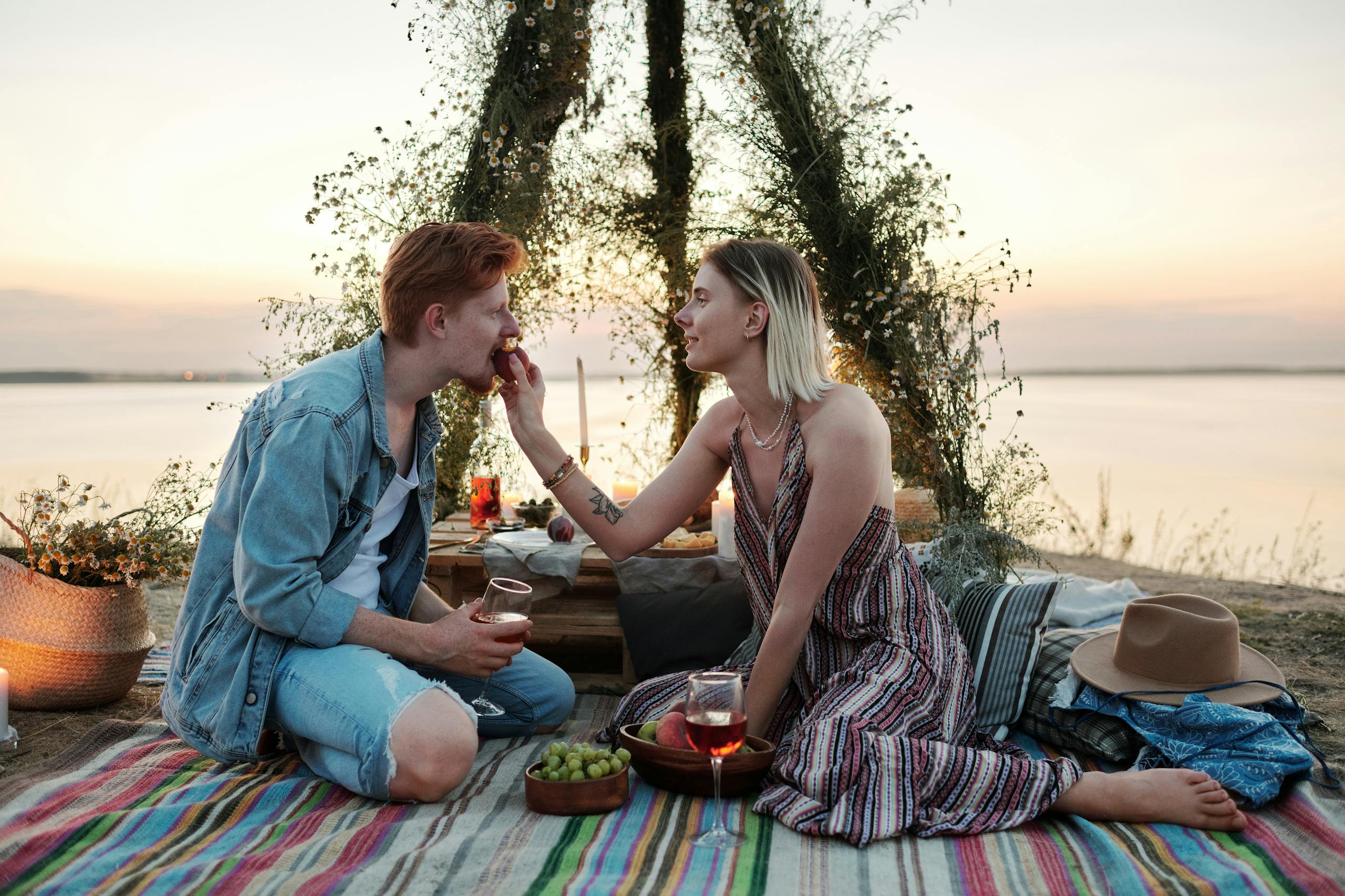 A couple enjoys a romantic picnic by the lakeside during sunset, sharing drinks and fruit.