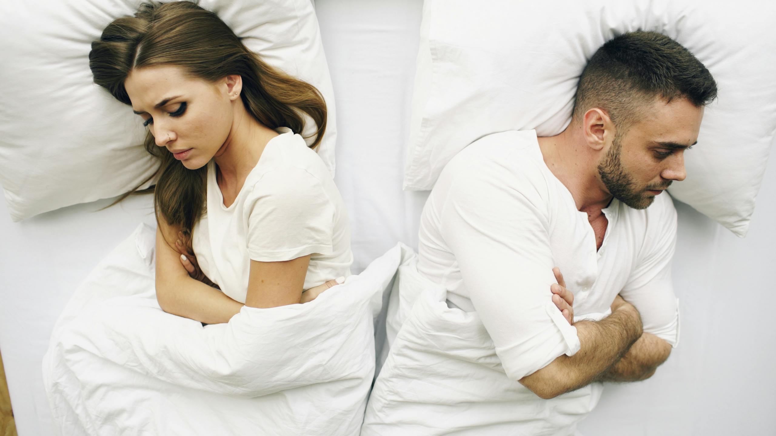A couple lying in bed facing away, expressions of discontent.