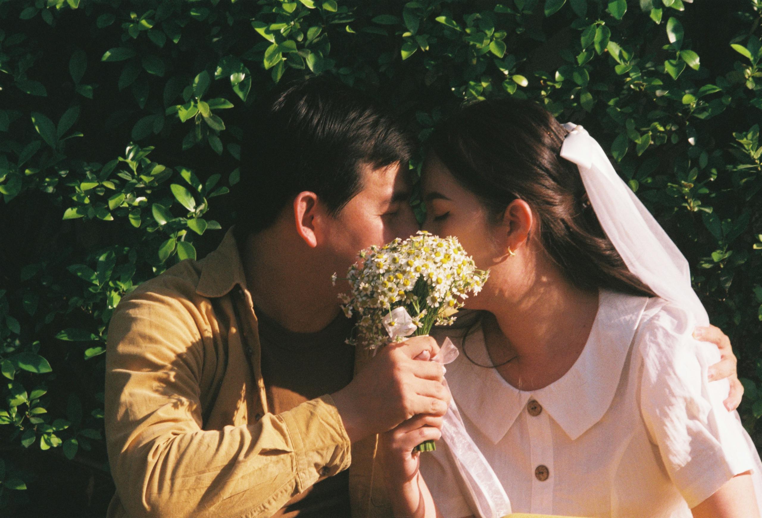 A couple sharing a tender moment, eyes closed, with a floral bouquet outdoors.