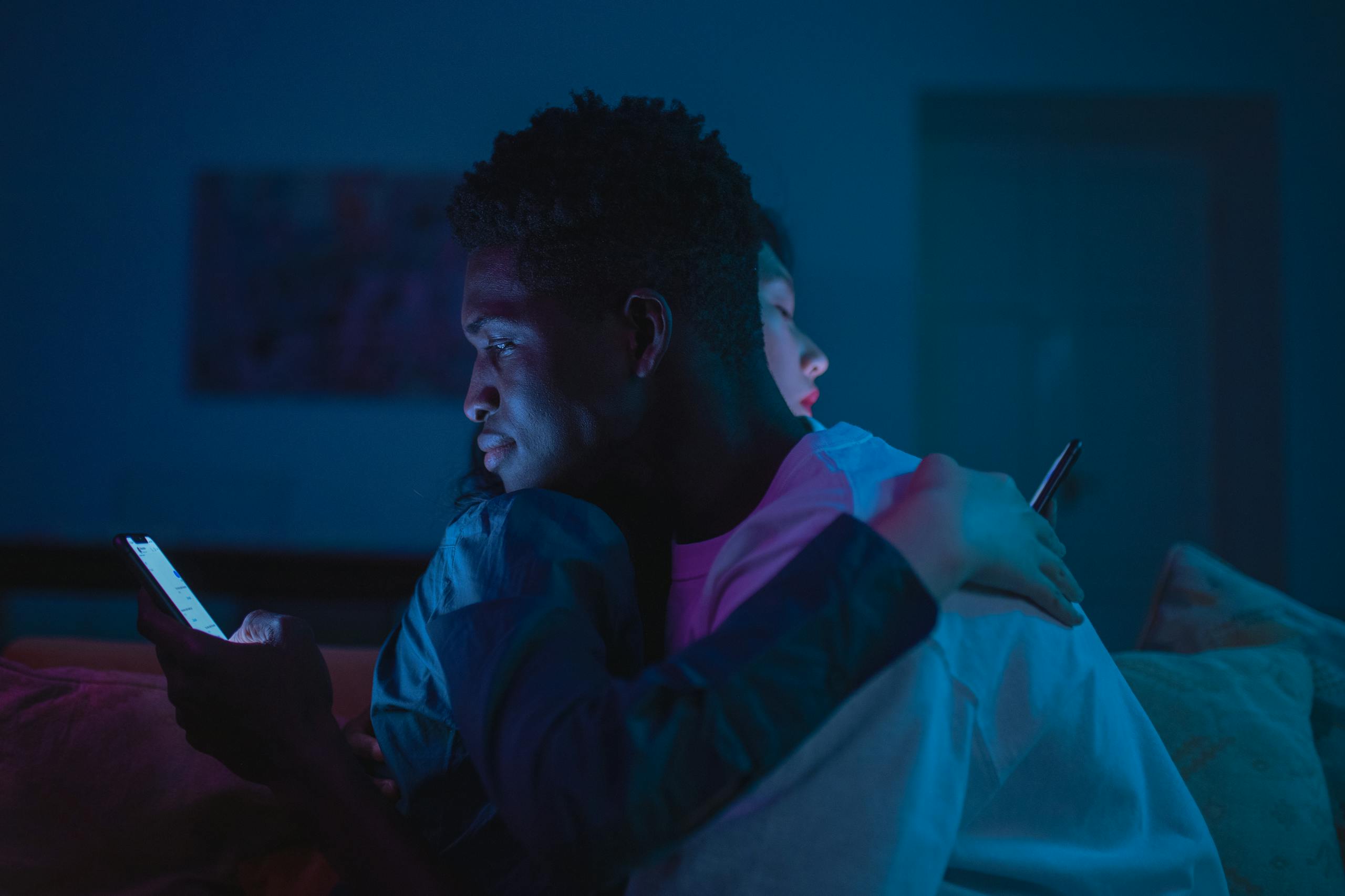 A couple sits closely in dim neon light, both absorbed in their smartphones.