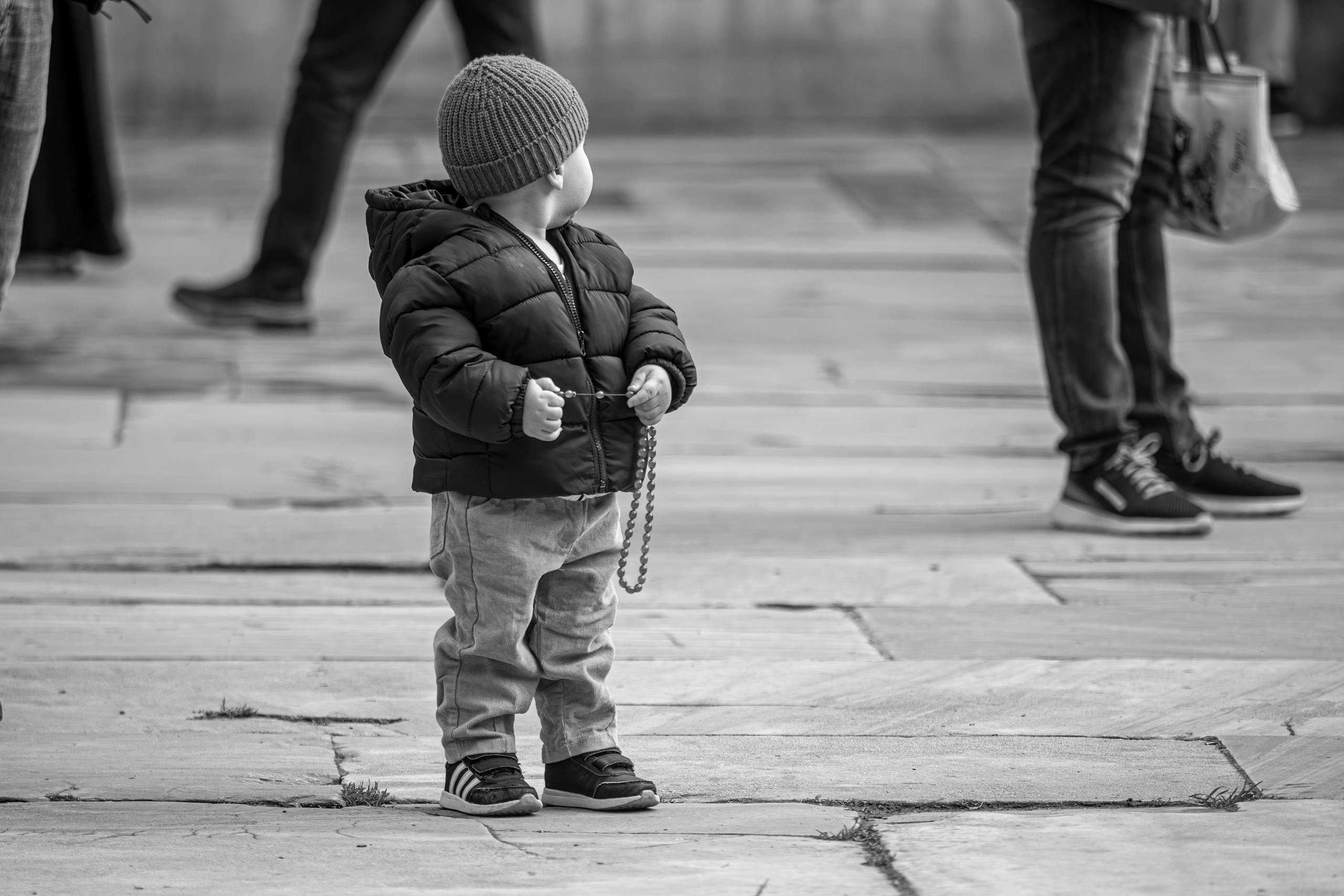 A cute toddler in warm clothing holding a string, surrounded by people in an outdoor setting.