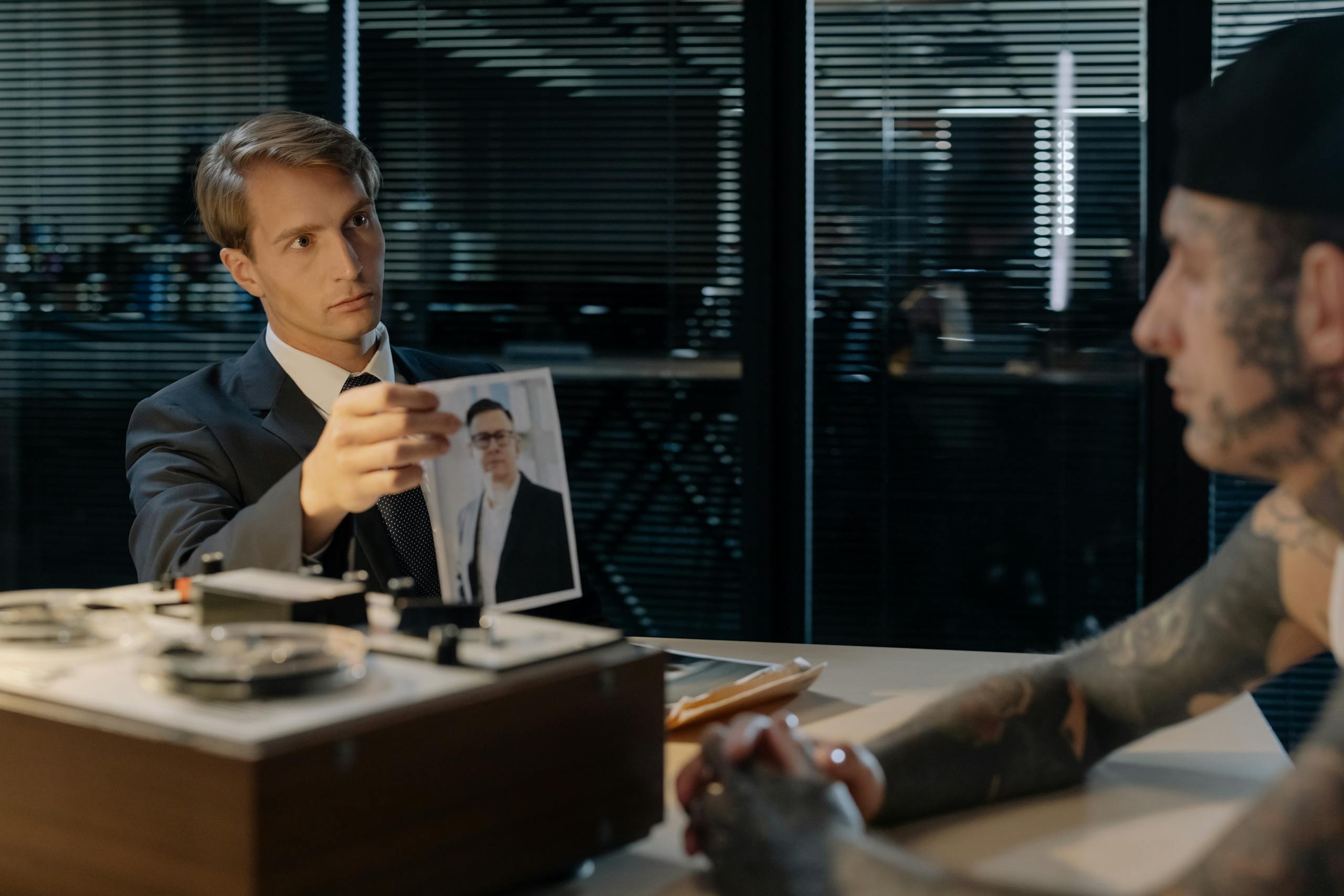 A detective interrogates a tattooed suspect in an office, examining a photograph.