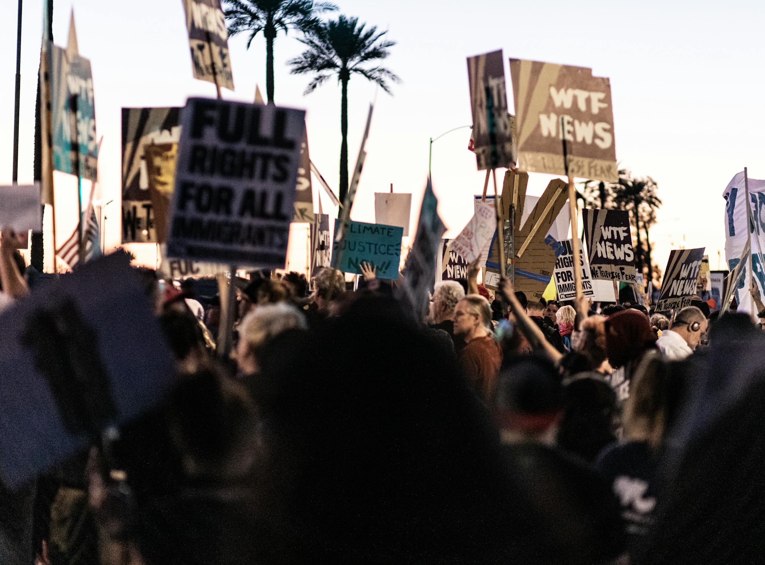 A diverse crowd gathers outdoors holding protest banners and signs at dusk.