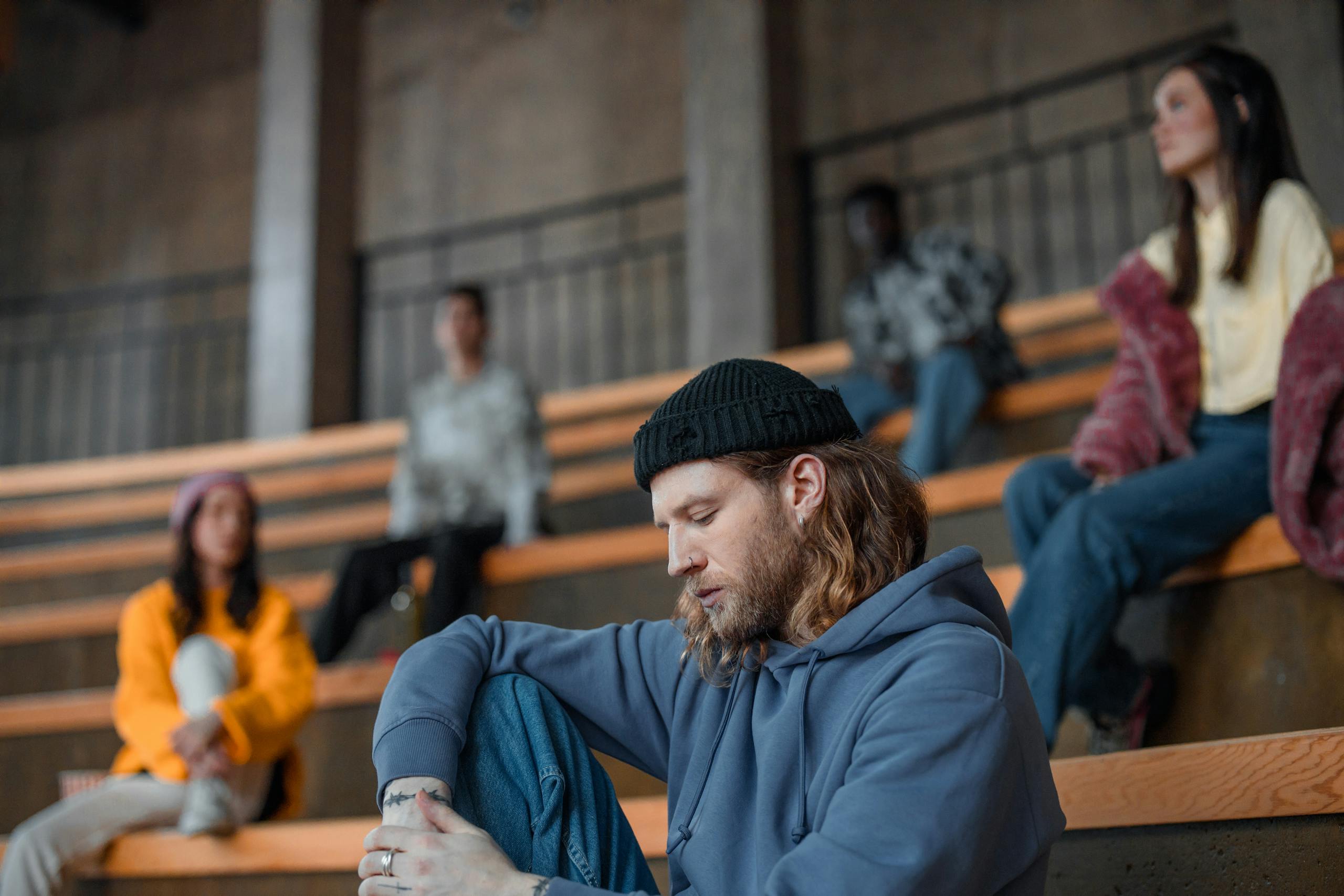 A diverse group of adults seated in an auditorium, focused on a pensive man in a hoodie and knitted cap.