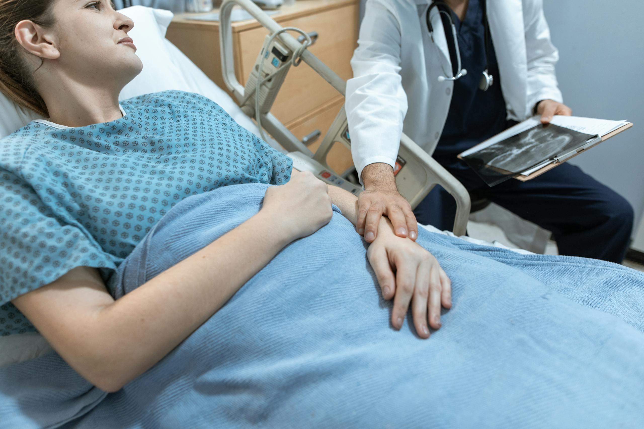 A doctor comforts a female patient in a hospital bed, holding her hand, with x-ray results.