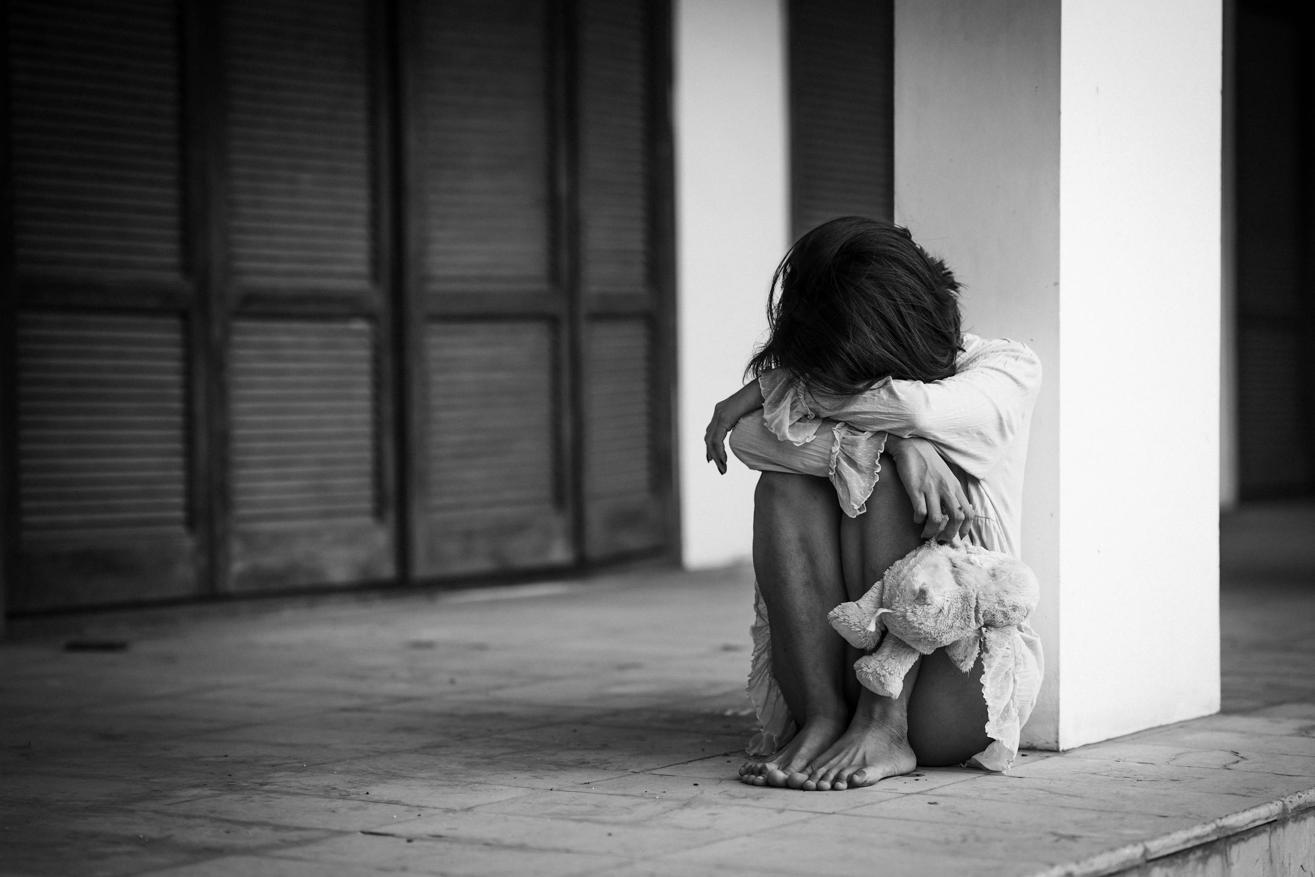 A dramatic black and white photograph of a woman sitting alone with a stuffed toy, evoking emotions of solitude and contemplation.