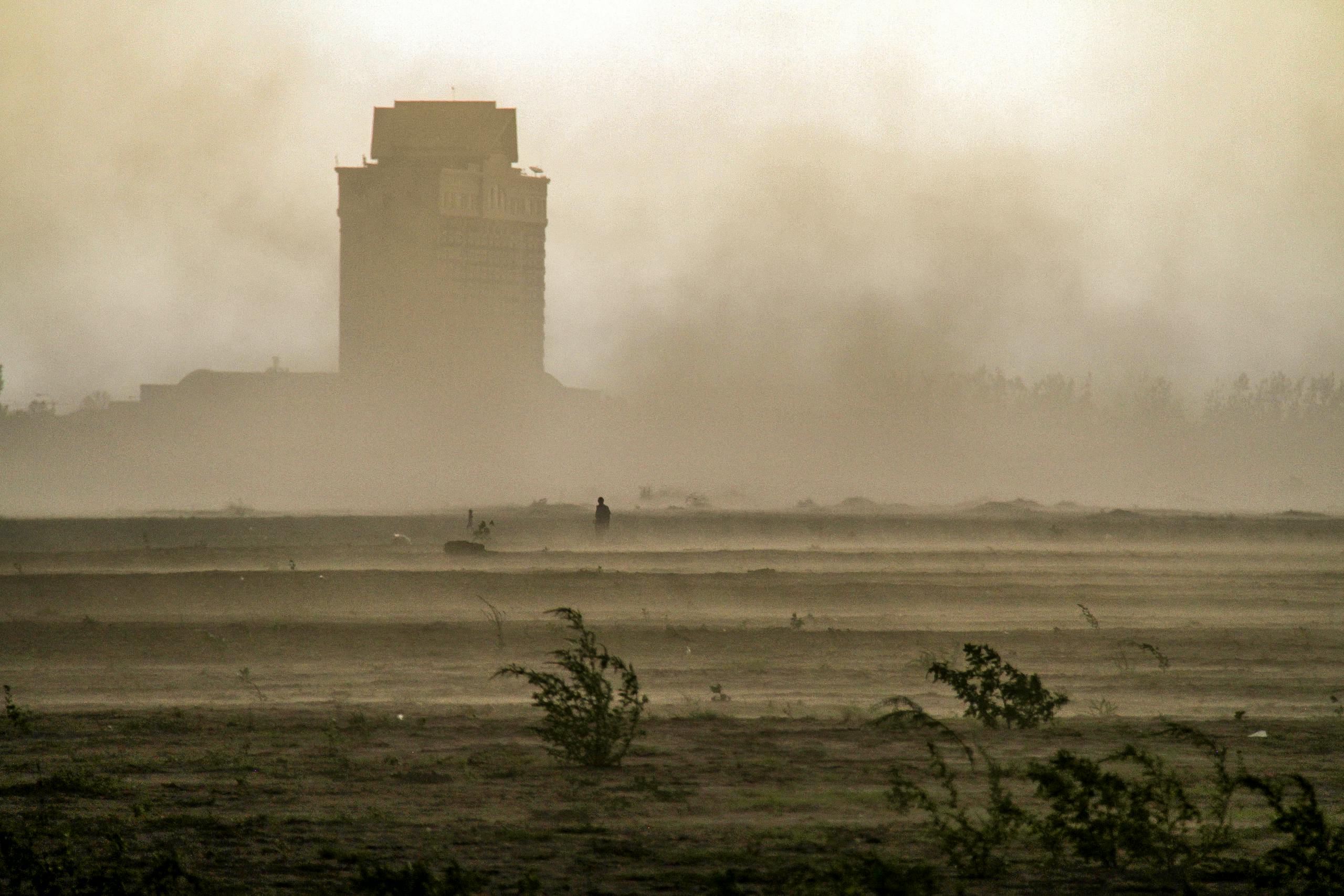 A dramatic scene of a dust storm engulfing a building and sparse vegetation in the foreground.