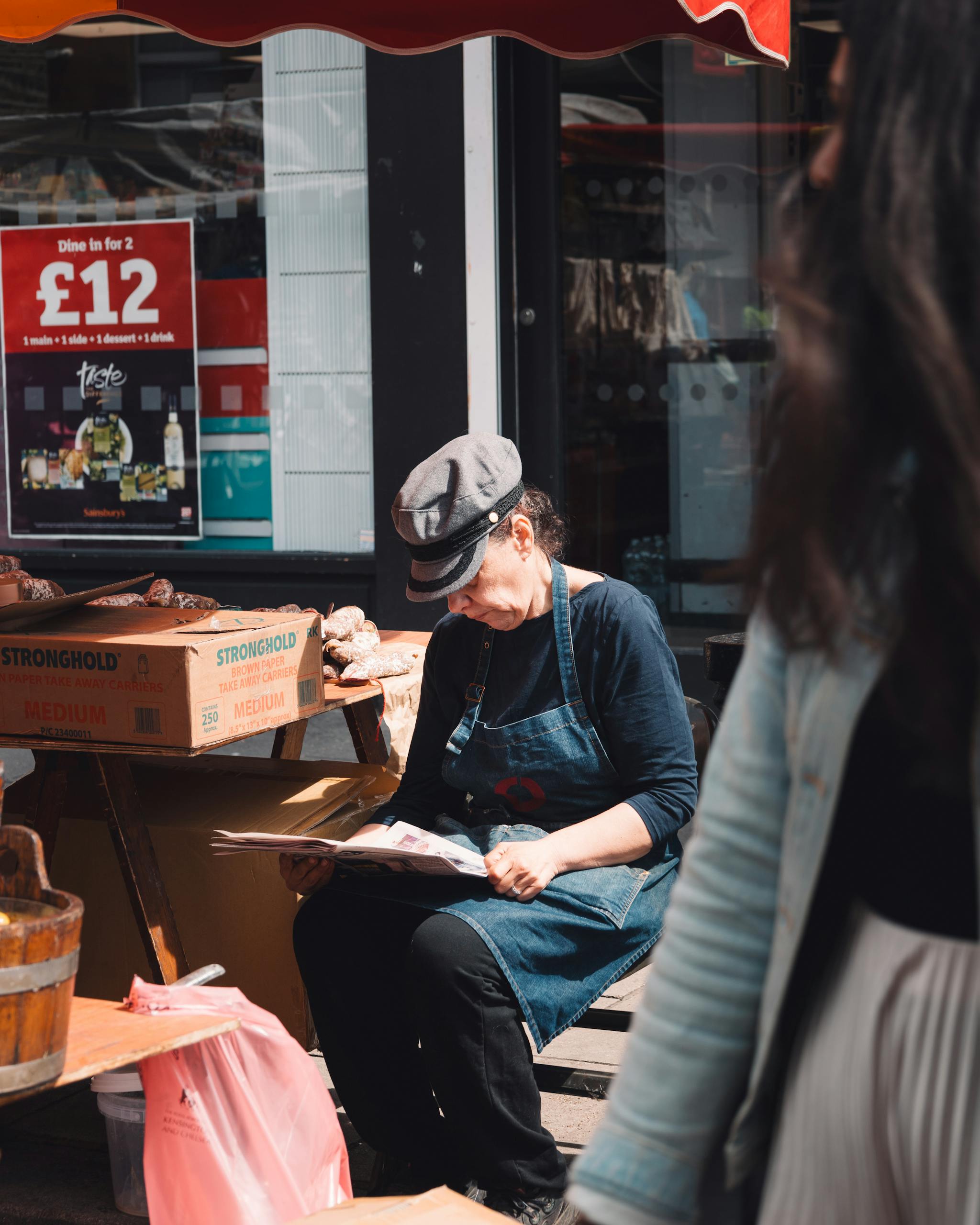 A female vendor reads a newspaper at a bustling outdoor market under a bright red awning.