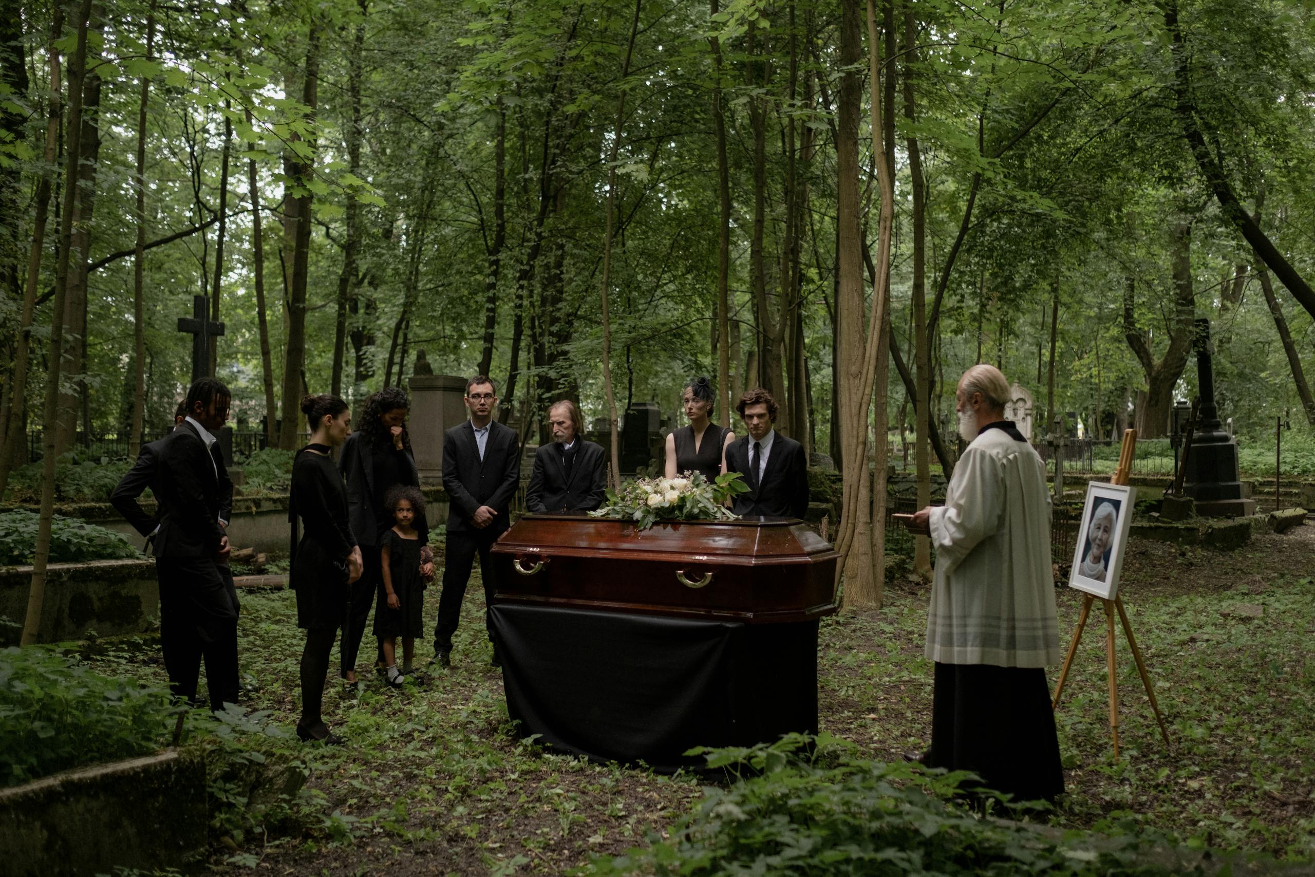 A group gathers at a solemn funeral in a forested cemetery, honoring the departed with a priest present.