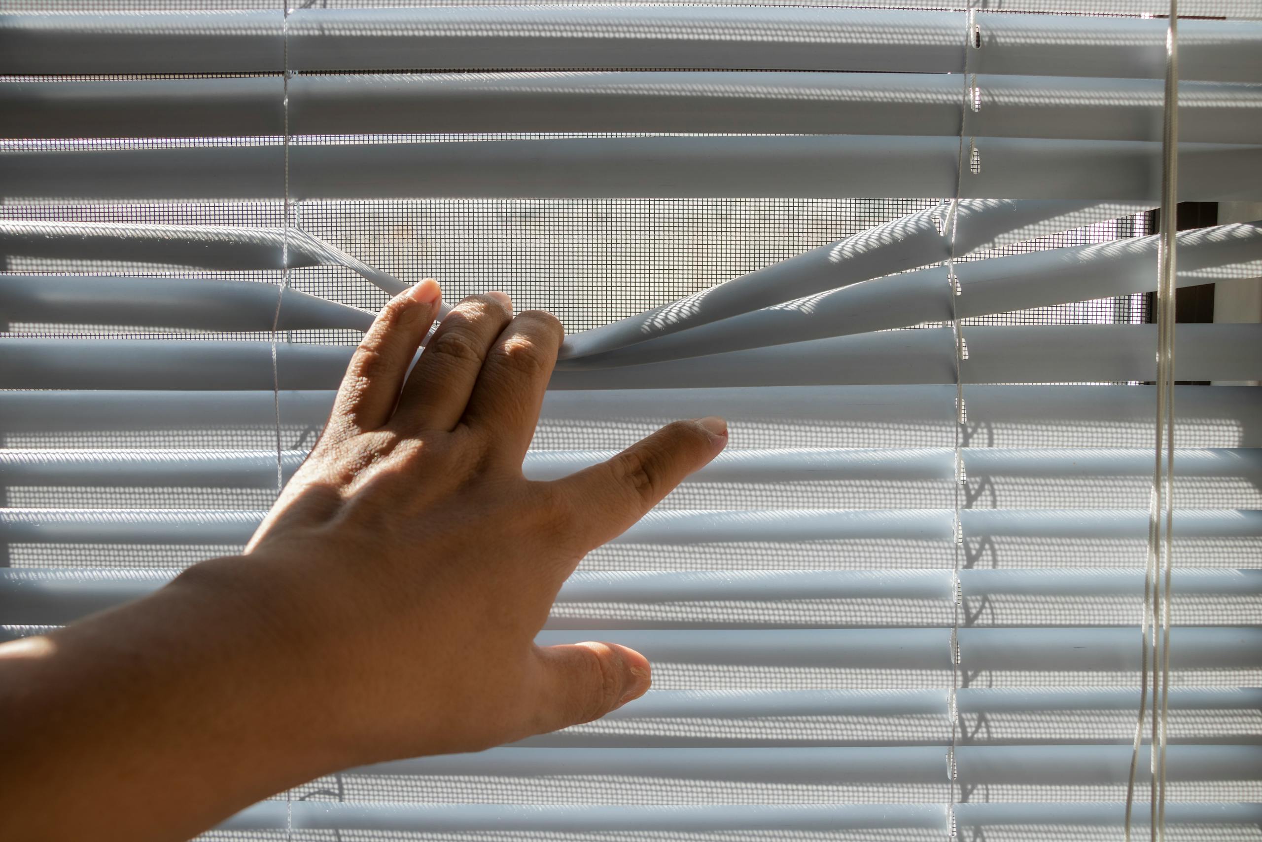 A hand gently opens window blinds, letting sunlight stream indoors.
