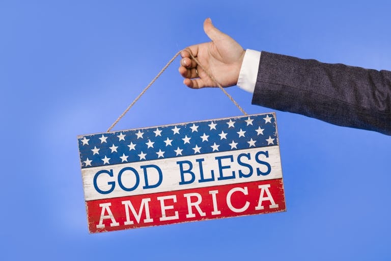 A hand holding a patriotic sign with 'God Bless America' text on a blue background.