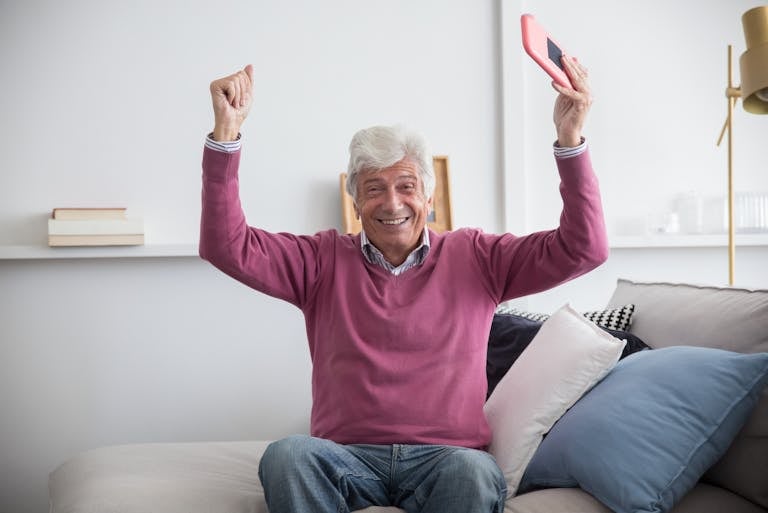 A happy senior man sitting on a sofa holding a smartphone indoors, celebrating with raised arms.