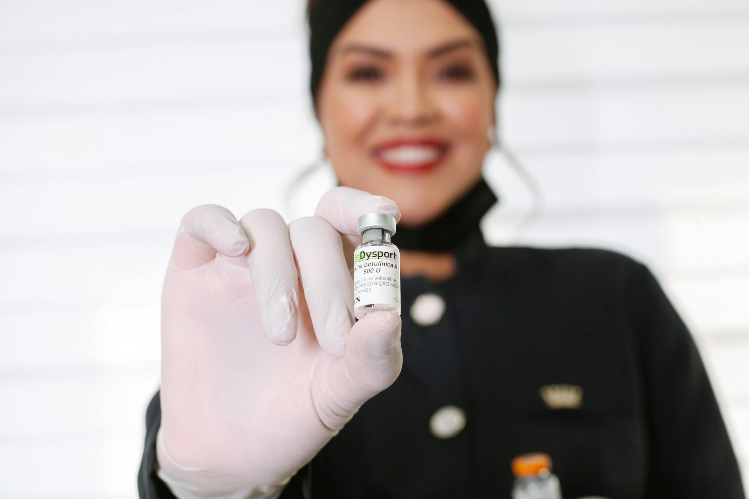 A healthcare worker in gloves holding a Dysport vial, smiling indoors.