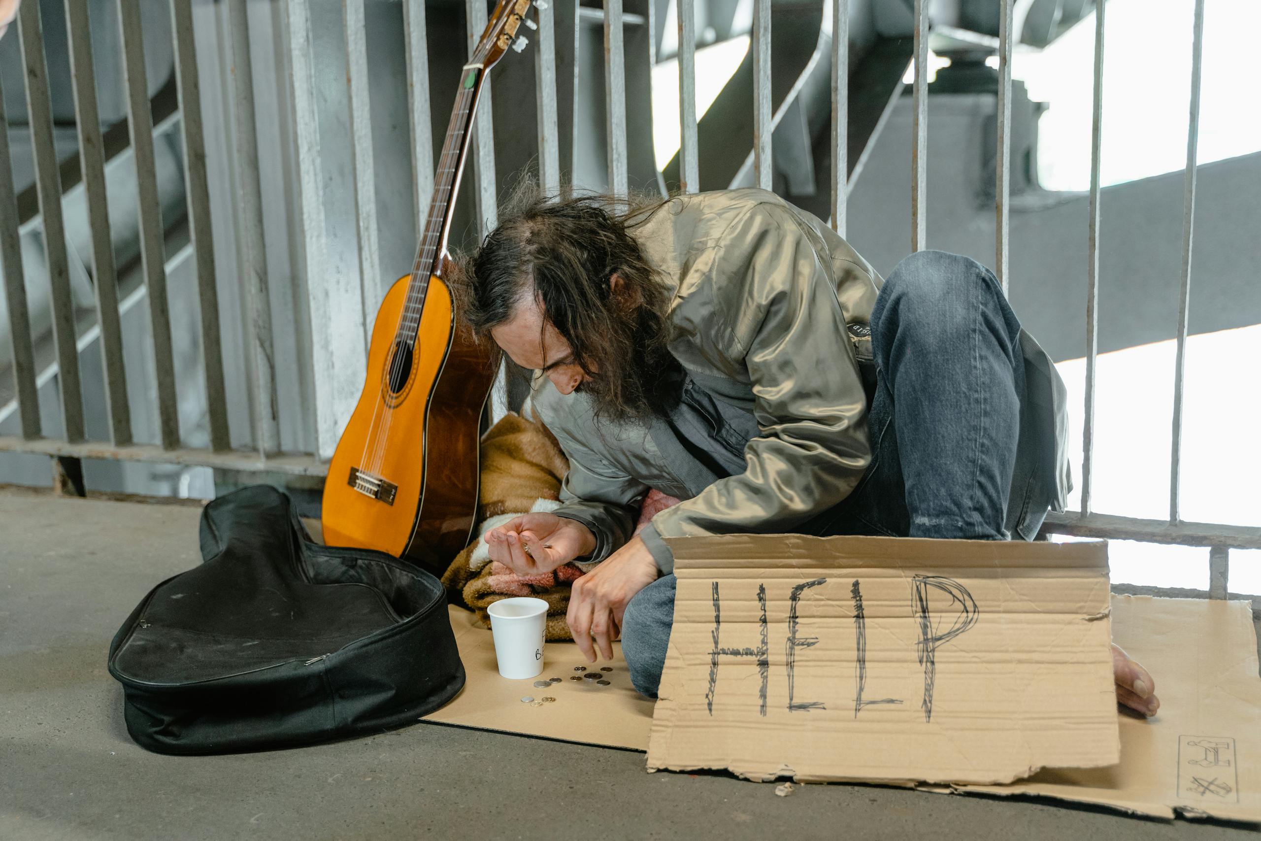A homeless man sits by a railing with a cardboard sign reading 'HELP', coins, and a guitar.