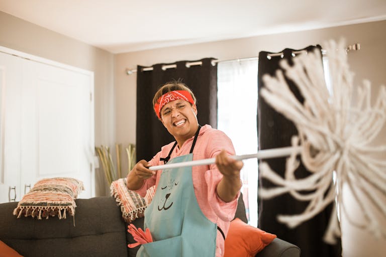A joyful man wearing an apron and bandana playfully holding a mop in a cozy living room.