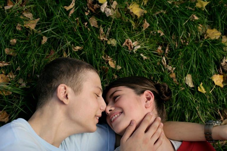 A joyful young couple smiles and embraces on grass with autumn leaves.