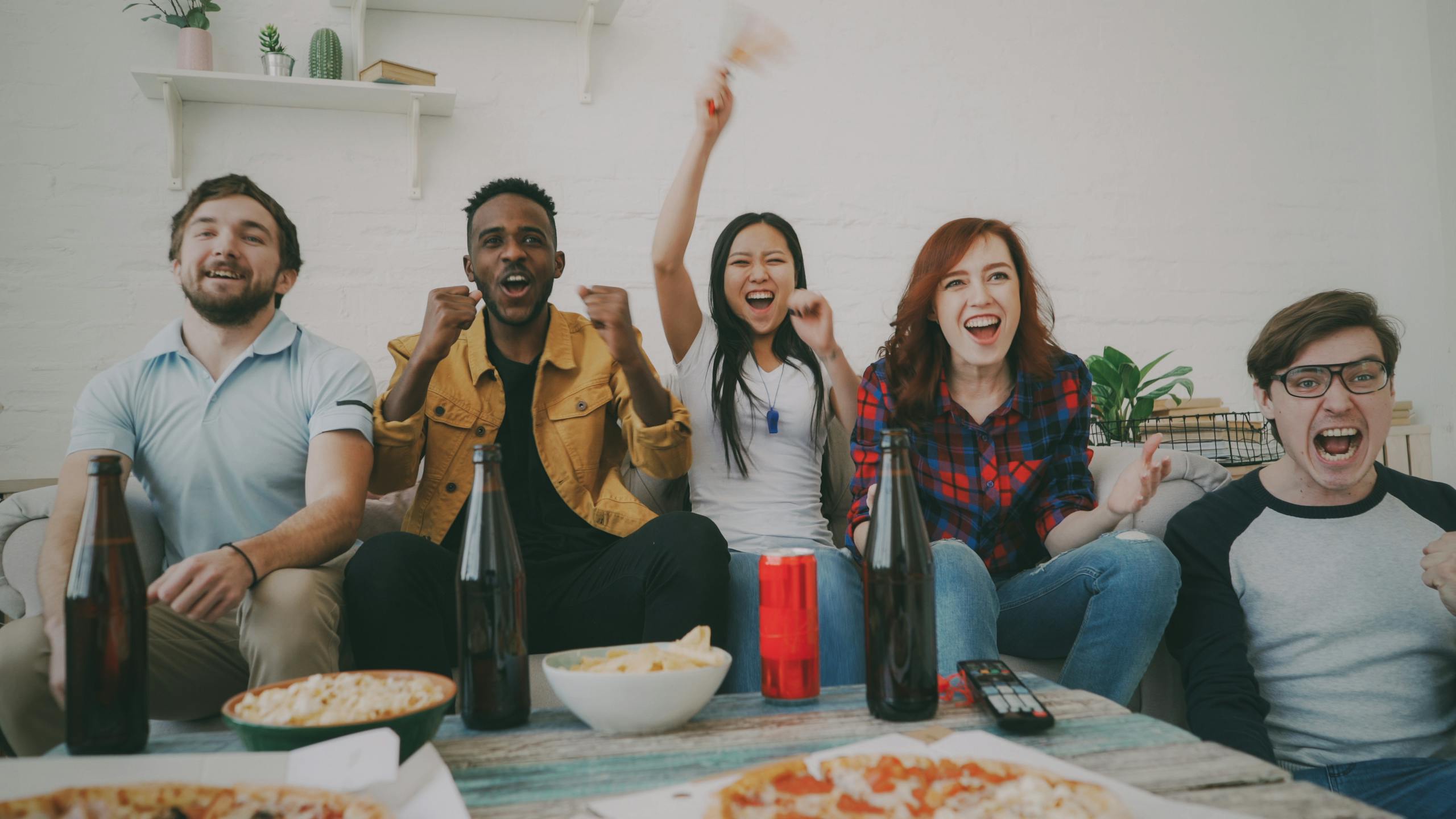 A lively group of friends cheering and watching a game indoors with snacks and drinks.