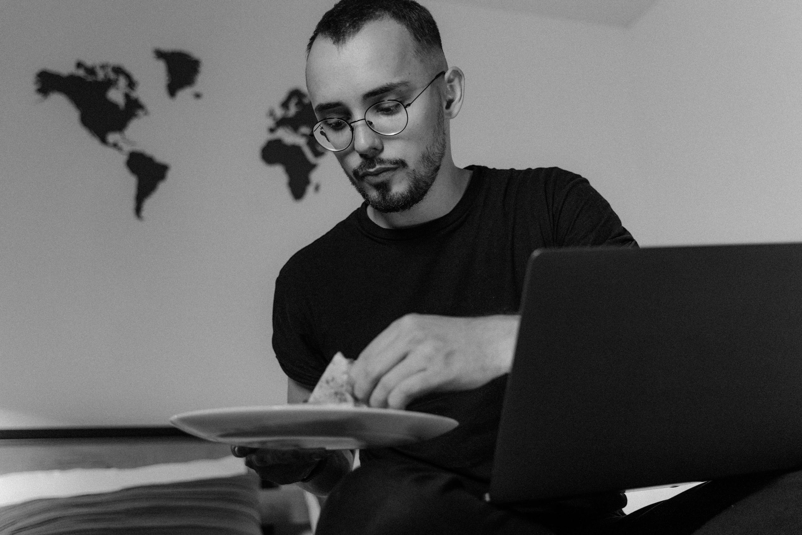 A man enjoys pizza while working remotely on his laptop in a cozy bedroom setting.