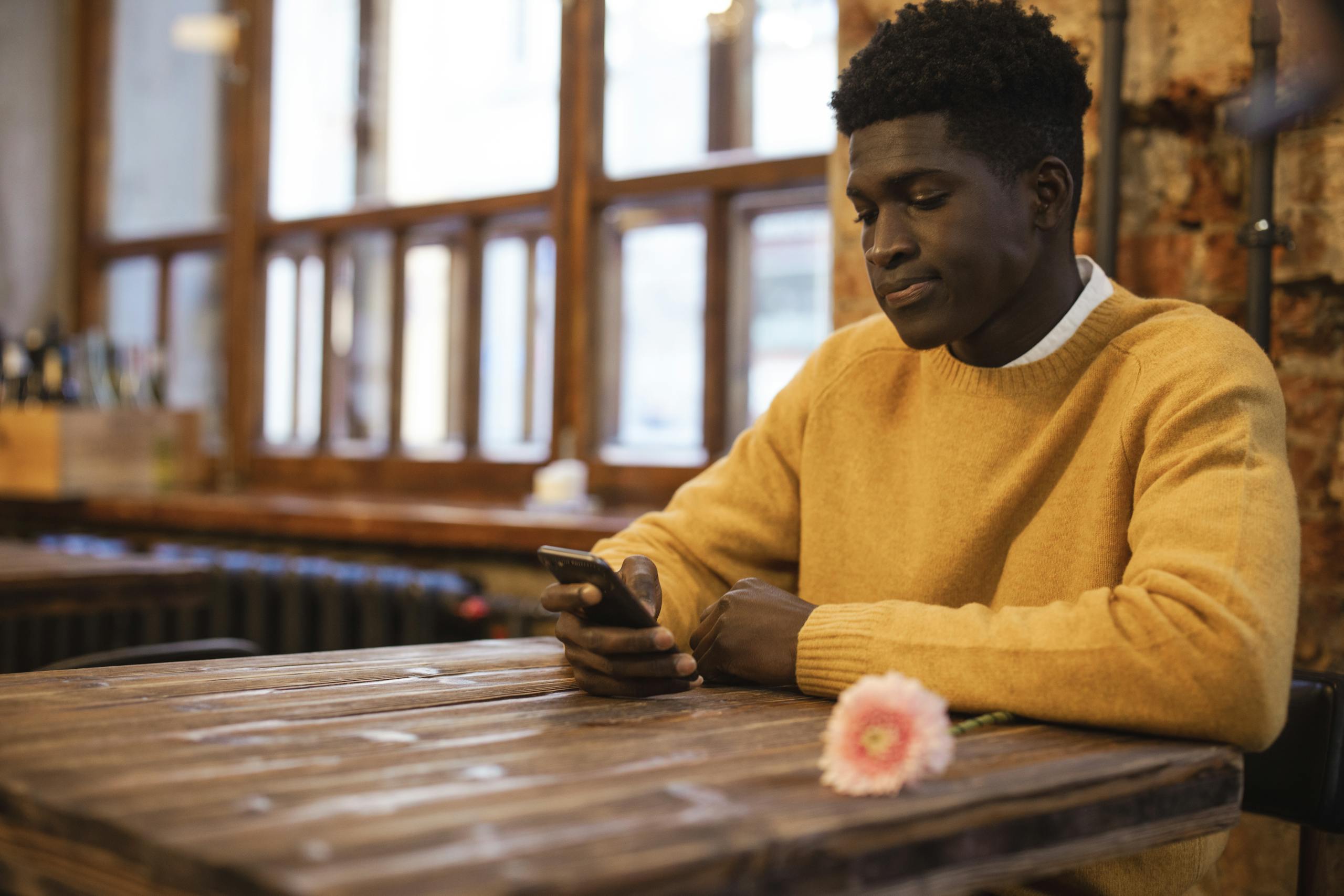 A man in a caf&eacute; using a smartphone, sitting at a wooden table with a flower, creating a warm and inviting atmosphere.