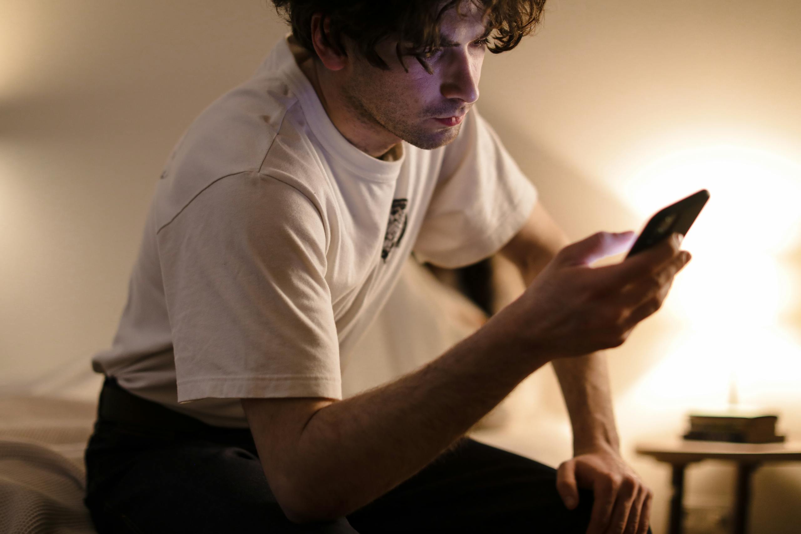 A man in a white shirt browses his smartphone indoors with warm lighting.