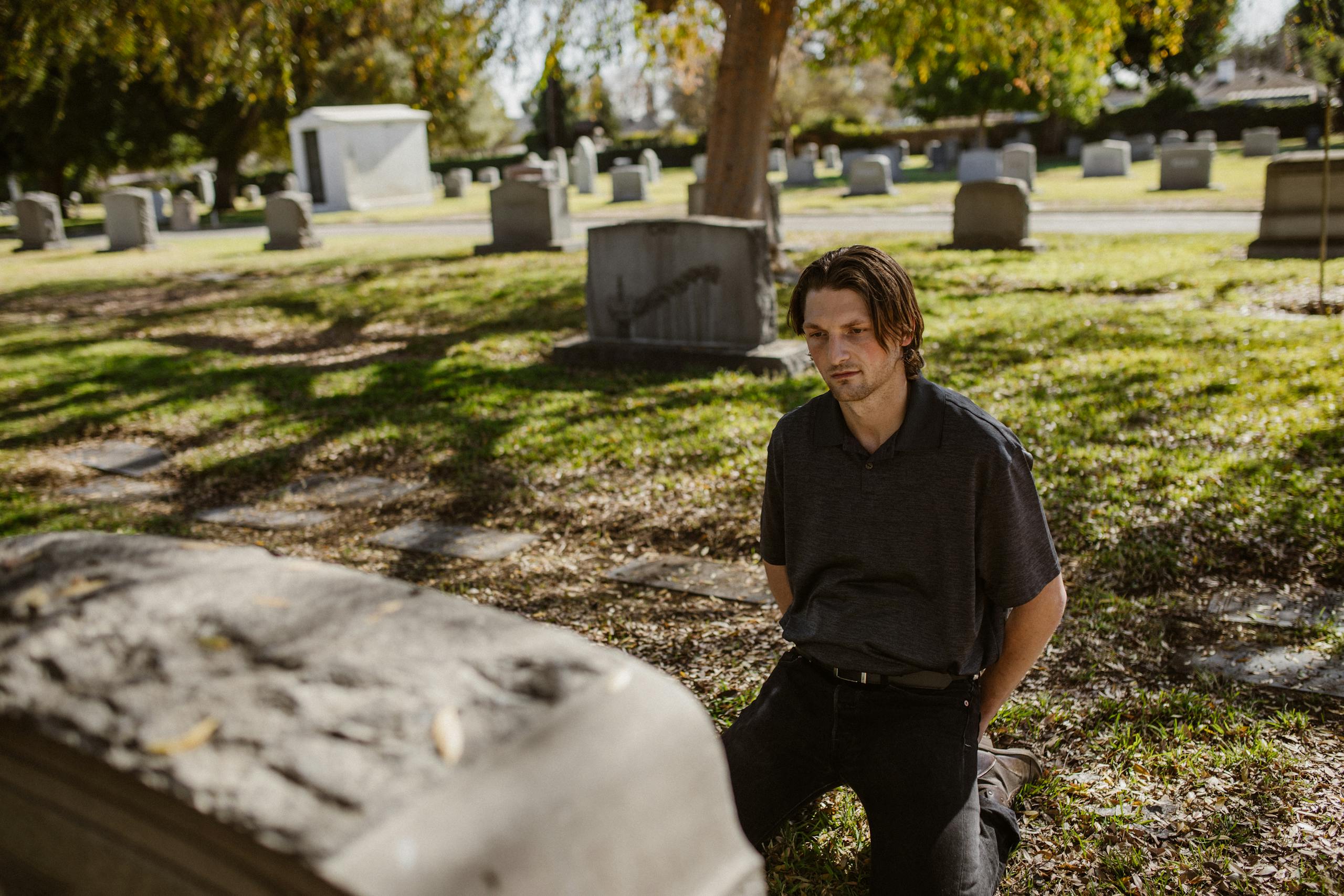 A man kneels at a grave in a sunlit cemetery, expressing grief and remembrance.