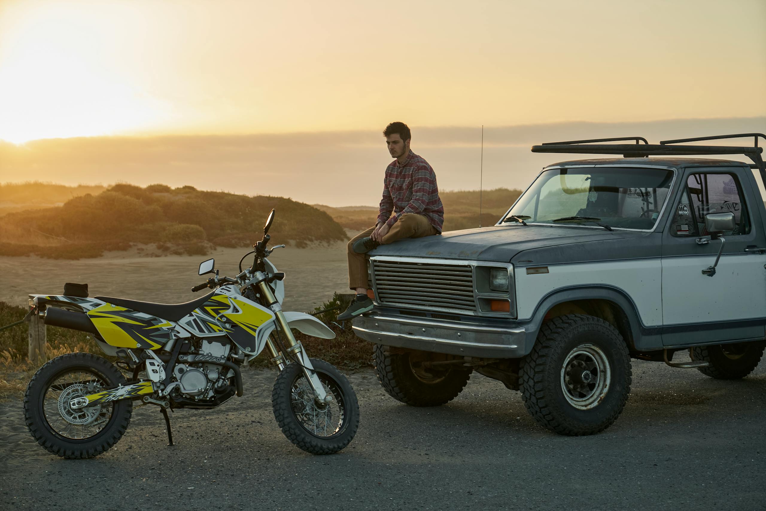 A man sits on a vintage truck by the seaside at sunset with a motorbike nearby.