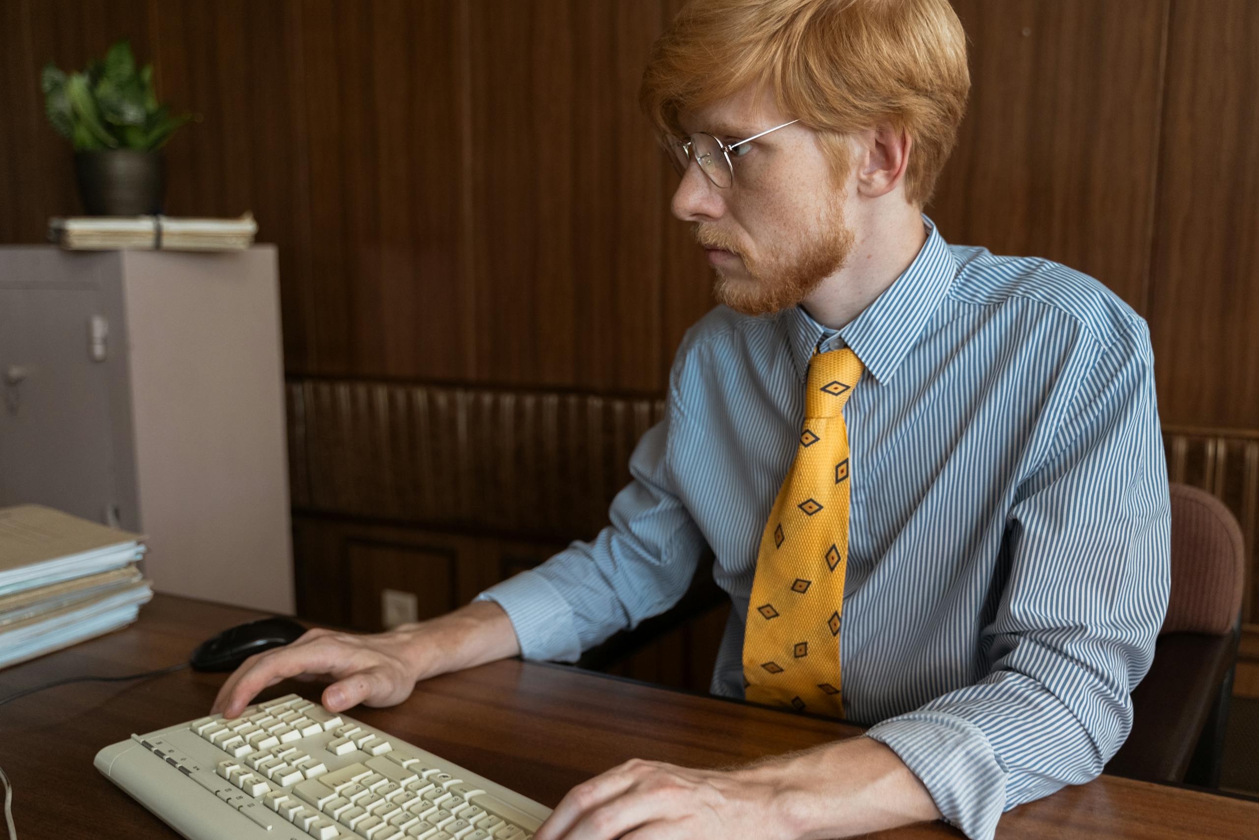 A man with glasses working at a desktop computer in an office setting.