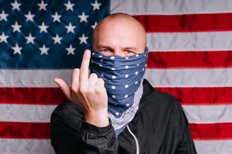 A masked individual making a hand gesture in front of an American flag, symbolizing protest.