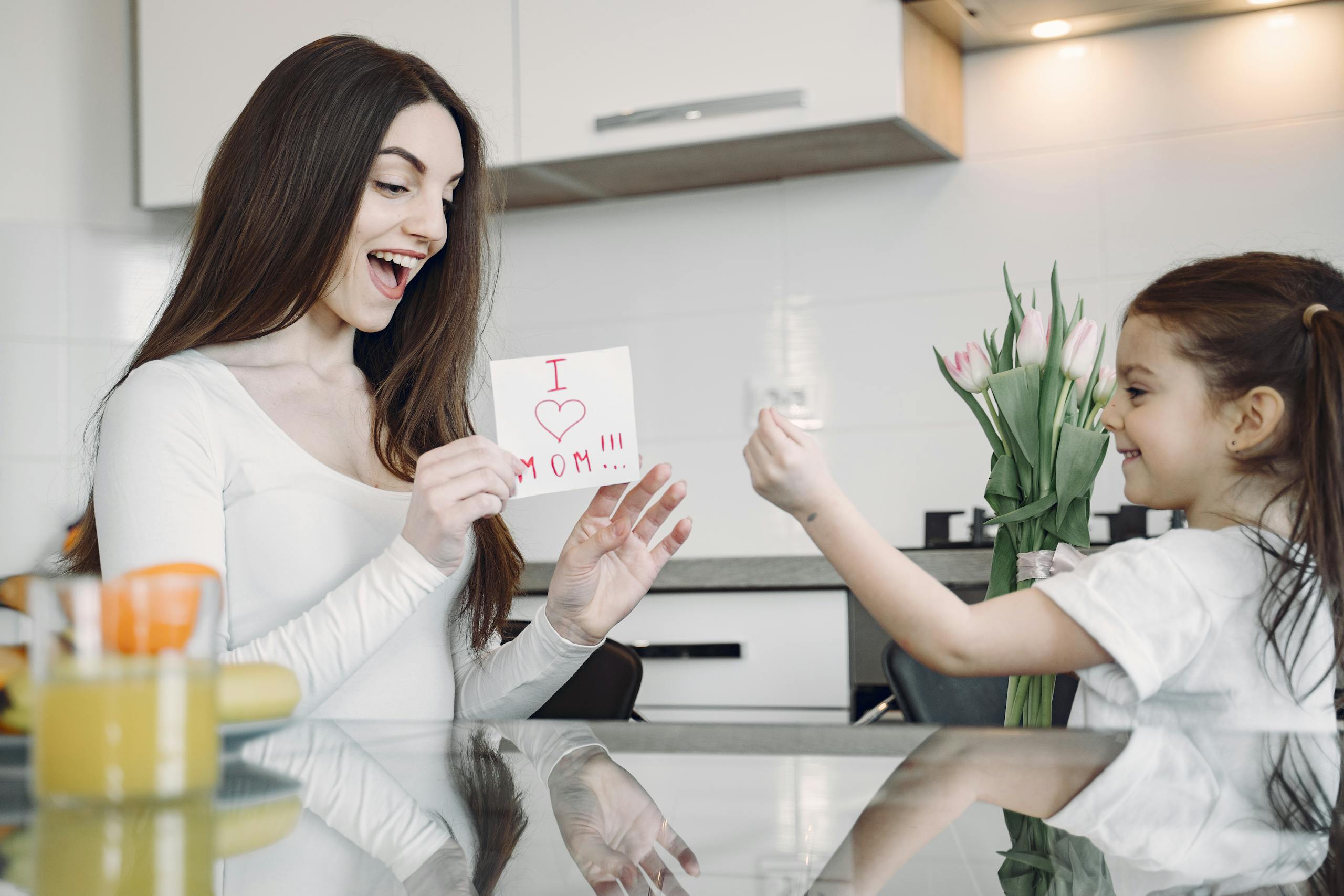 A mother and daughter sharing a joyful moment with flowers and a card in a kitchen.