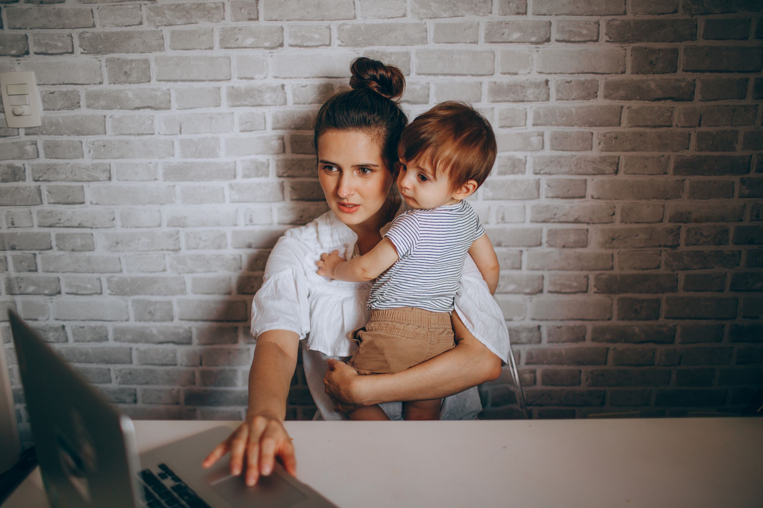 A mother multitasks by working on her laptop while holding her baby in a cozy home environment.