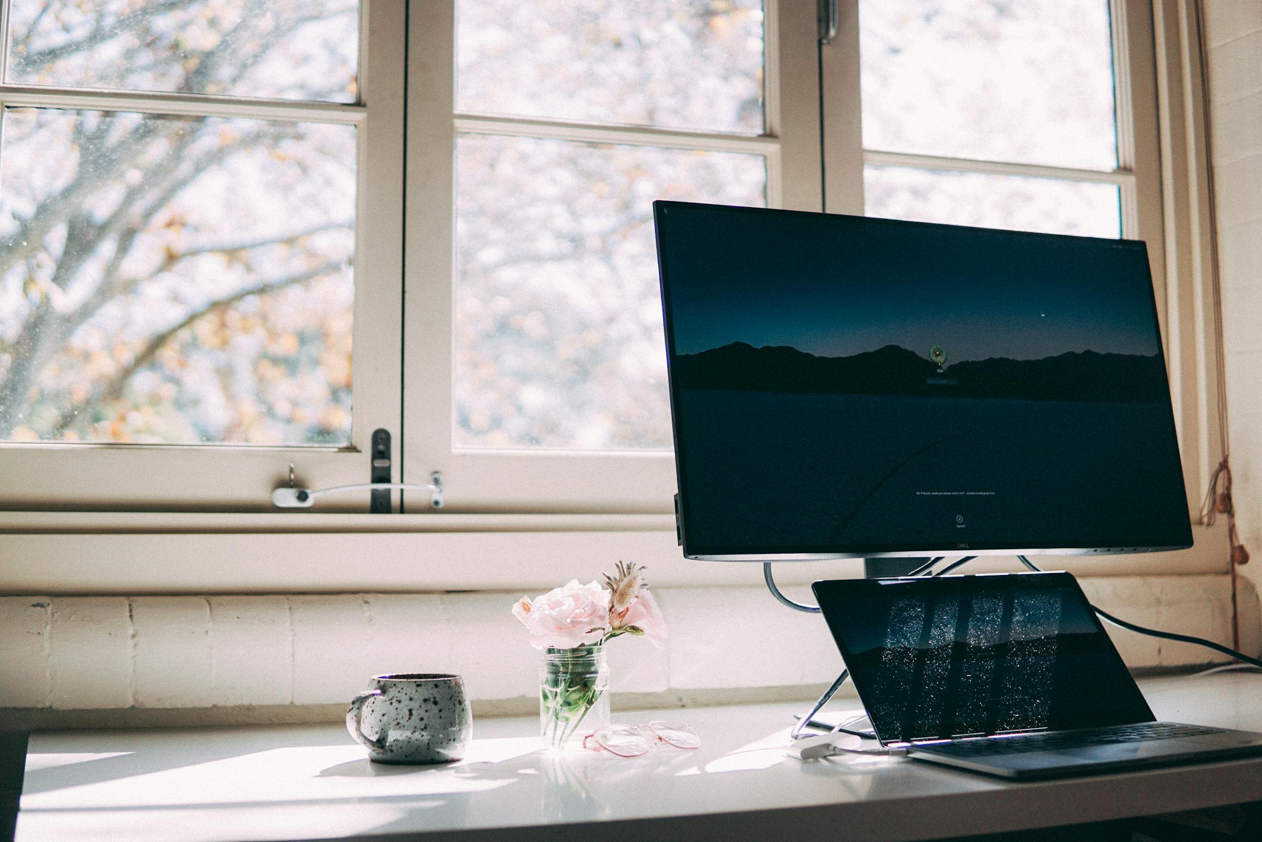 A peaceful home office setup with a desktop and laptop by a window, decorated with flowers and a mug.
