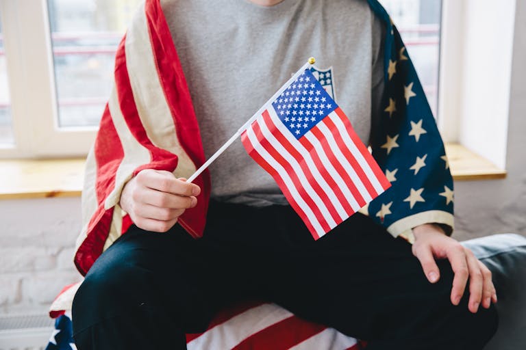 A person sitting indoors, holding a small American flag, draped in a larger flag, celebrating Independence Day.
