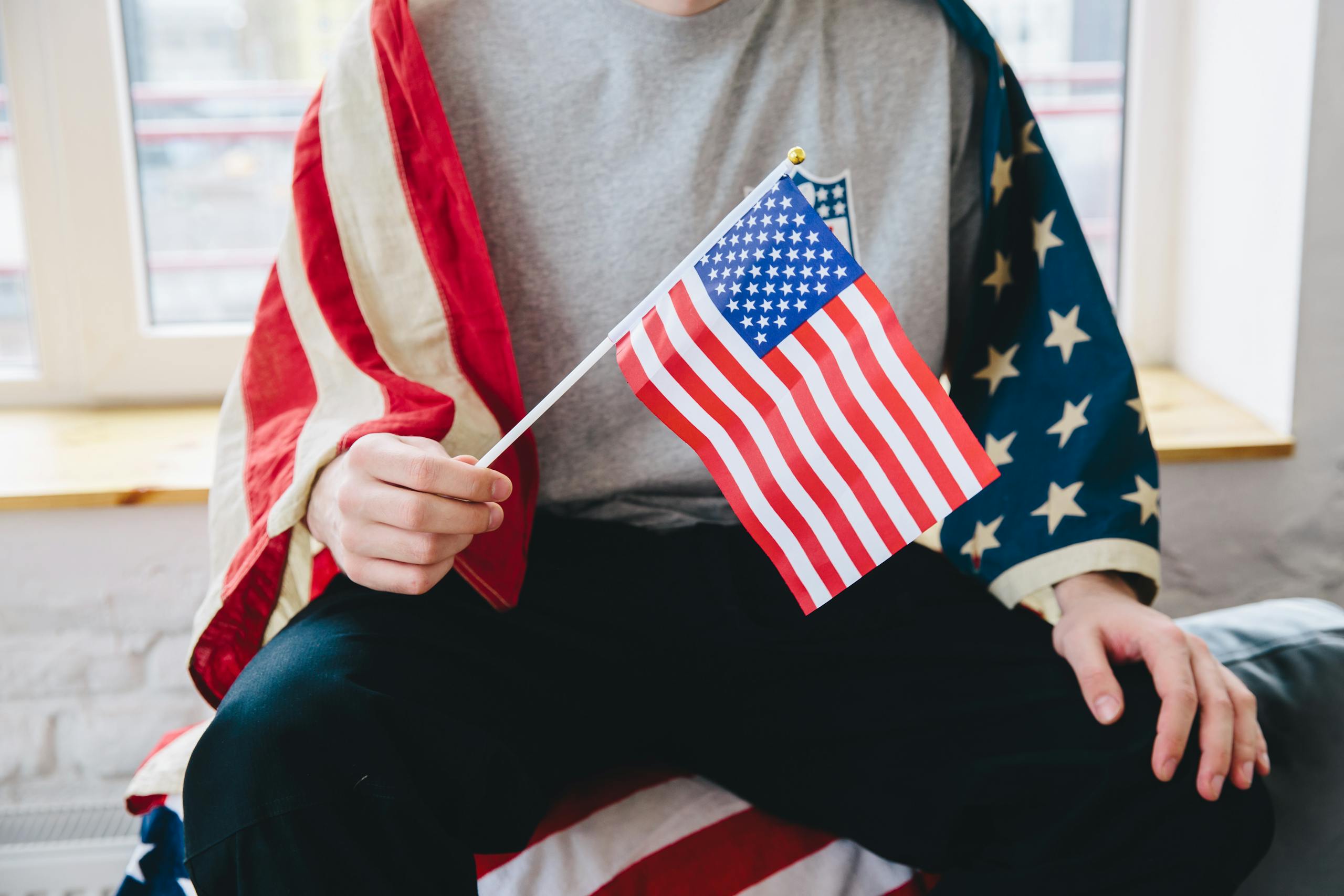 A person sitting indoors, holding a small American flag, draped in a larger flag, celebrating Independence Day.