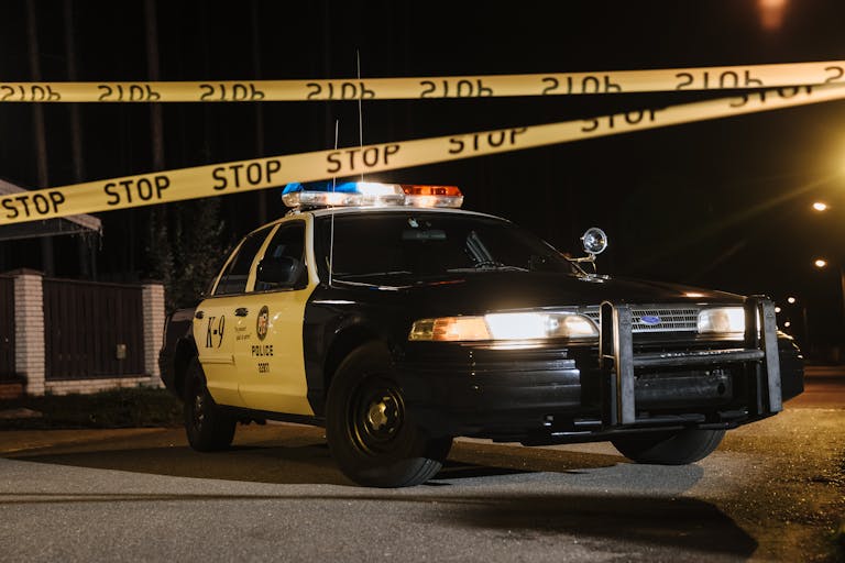A police car parked at a night crime scene with yellow barricade tape, under streetlights.