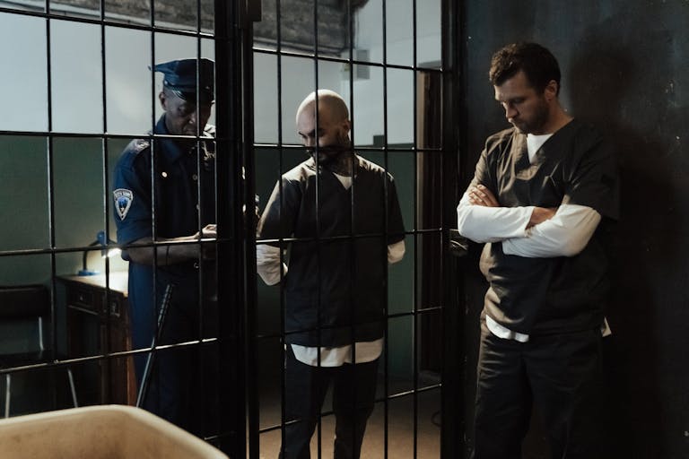 A police officer stands by two prisoners in jail uniforms behind bars, indoors.