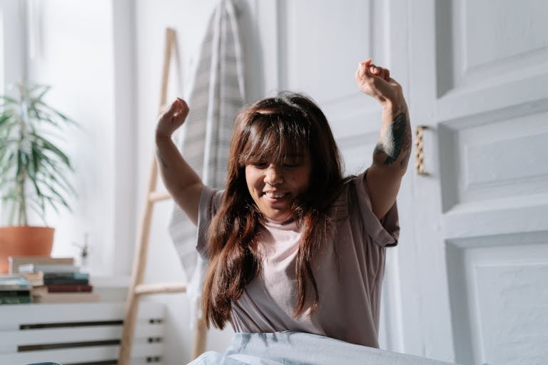 A relaxed Asian woman wakes up and stretches in a sunlit bedroom, enjoying the morning light.