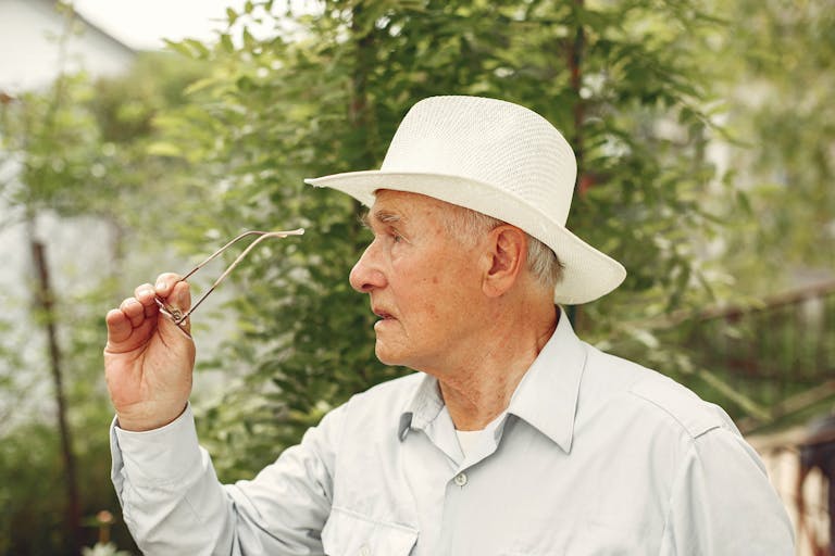 A senior man in a white hat holds his glasses thoughtfully in an outdoor setting. Summer vibes.