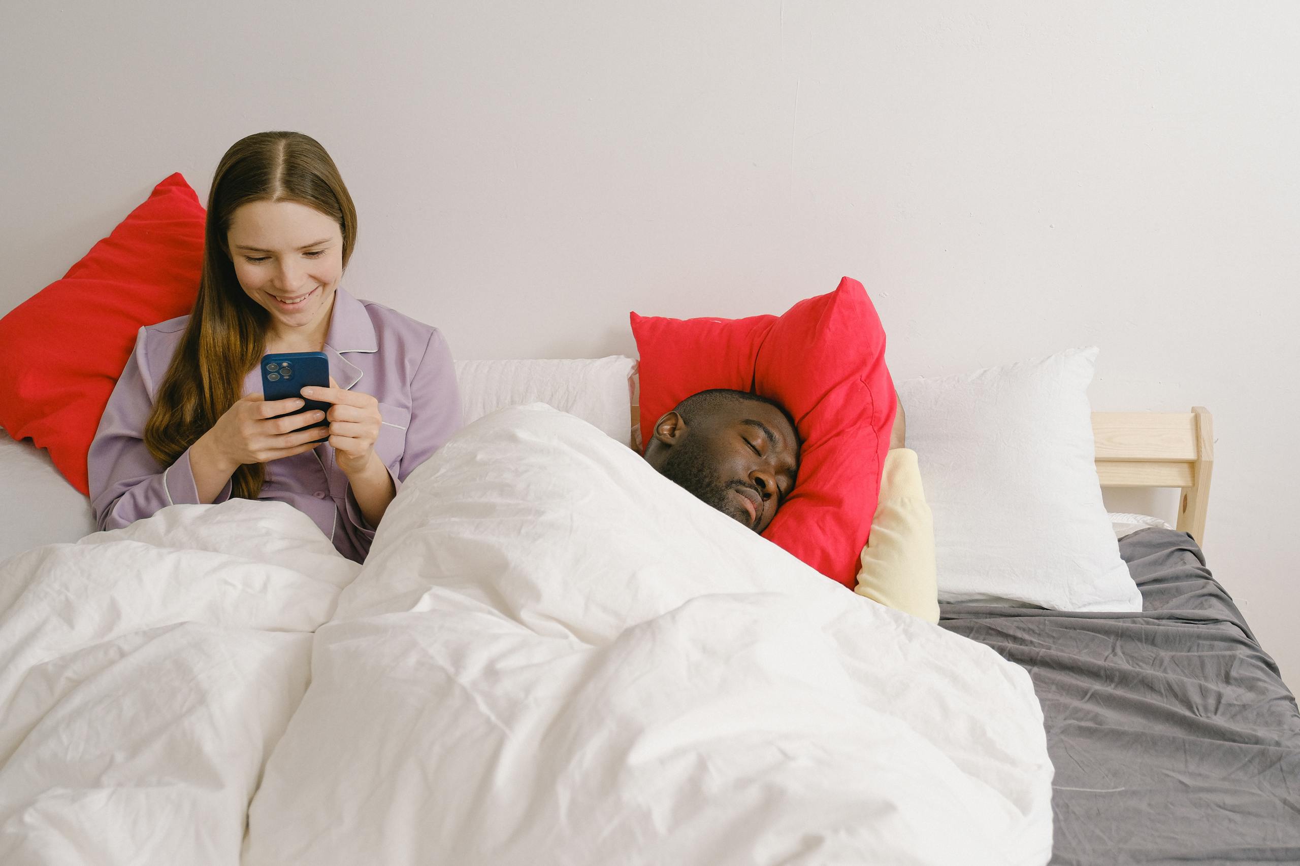 A smiling woman on her phone next to a sleeping man in a cozy bedroom setting.